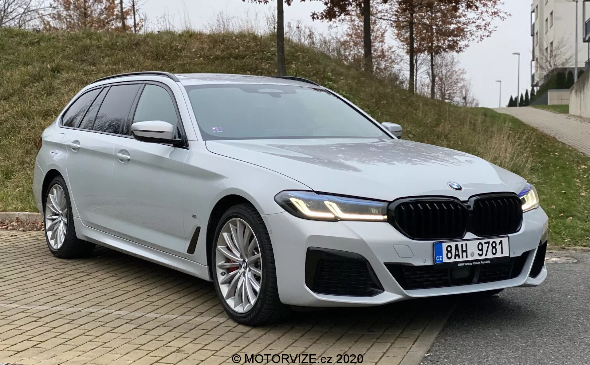 Egy modern, luxus BMW 5-ös széria (BMW 520i Touring Steptronic) kombi alacsony, az elülső háromnegyedes szögből fényképezve. A kép kiemeli az autó elülső hűtőrácsát, a valószínűleg LED technológiát használó, elegáns, szögletes fényszórókat, valamint a motorháztetőn és a sárvédőkön található izmos dizájnvonalakat. A látható részek közé tartozik az elülső hűtőrács, lökhárító, fényszórók, a sofőr oldalán található első ajtó, első sárvédő, a hátsó ajtó egy része, oldalsó ablakok, kerekek és a sofőr oldalán lévő oldalsó tükör. A nagy, ötvözött kerekek egy sportosabb változatra utalnak, és az ezüst festés visszaverő és tiszta. Az autó jellegzetes függőleges rácsokkal ellátott elülső hűtőrácsot, a magasabb felszereltségi szintet jelző fender emblémát és beépített irányjelzőkkel ellátott oldalsó tükröket tartalmaz. Egy napfénytető is látható. A jármű egy városi út szélén parkol, ahol az előtérben járda és padka látható, a háttérben pedig füves terület és egy épület, borús fényviszonyok között.