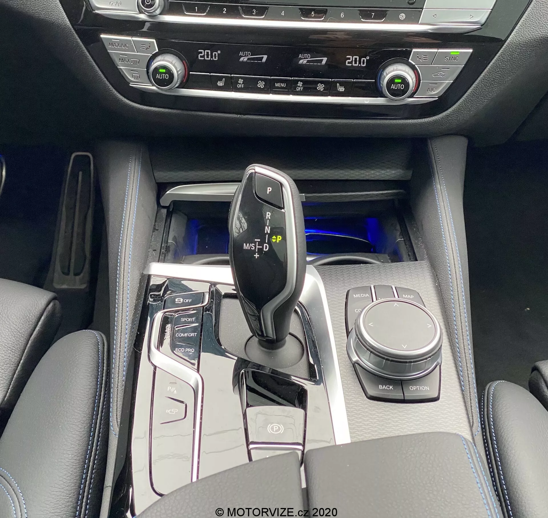 Madaras nézet a BMW 520i Touring Steptronic 2020 középkonzoljáról, amelyen látható a fényes fekete sebességváltó kar fémes díszítésekkel, körülötte pedig magas fényű kezelőpanel gombokkal a start/stop rendszerhez, vezetési módokhoz, parkolószenzorokhoz és máshoz. Látható az iDrive vezérlőtárcsa, pohártartók és párnázott kartámasz tárolóval. A hideg kék hangulatvilágítás fokozza a luxus érzetét. A kétzónás HVAC és ülésfűtés vezérlők fejlett kényelmi funkciókat jeleznek. A belső anyagok bőrhatású felületeket, fémes díszítéseket és magas fényű paneleket tartalmaznak.