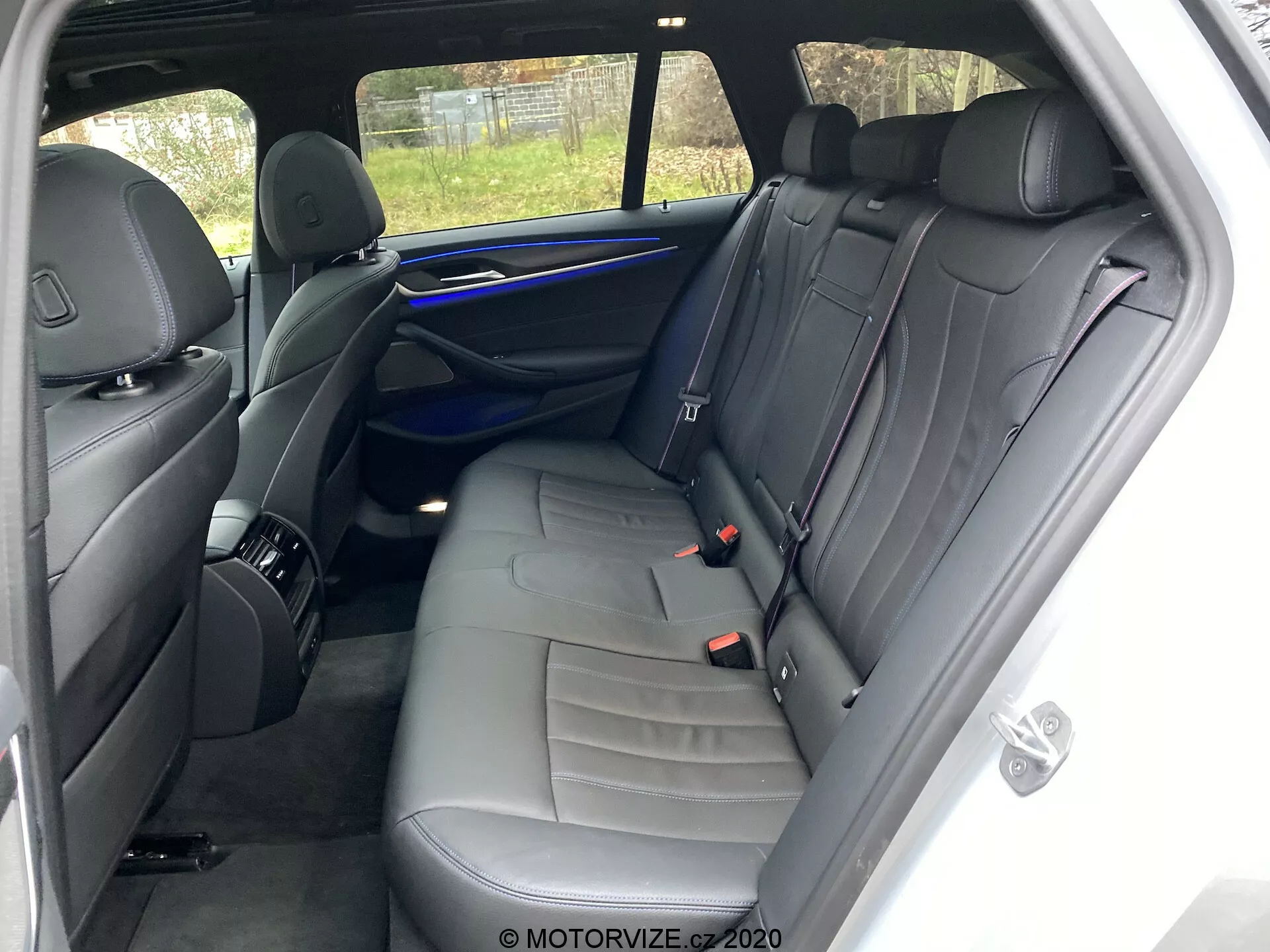 A 2020-as BMW 520i Touring Steptronic belső tere, a hátsó ülések területére fókuszálva a jobb oldalról. A nézőpont a jobb hátsó utasajtó közelében van, megmutatva egy háromszemélyes padot sötétbőr kárpitozással és díszvarrással. A funkciók közé tartozik három fejtámla, három biztonsági öv, egy ajtó panel hangszóró ráccsal, fogantyúval és vezérlőelemekkel, kék környezeti világítási szalag és tárolóháló az első utasülés hátoldalán. A jól karbantartott belső térben nincs látható személyes tárgy vagy jelentős kopás. A hátsó ablakon keresztüli részleges kilátás egy kerítést mutat.