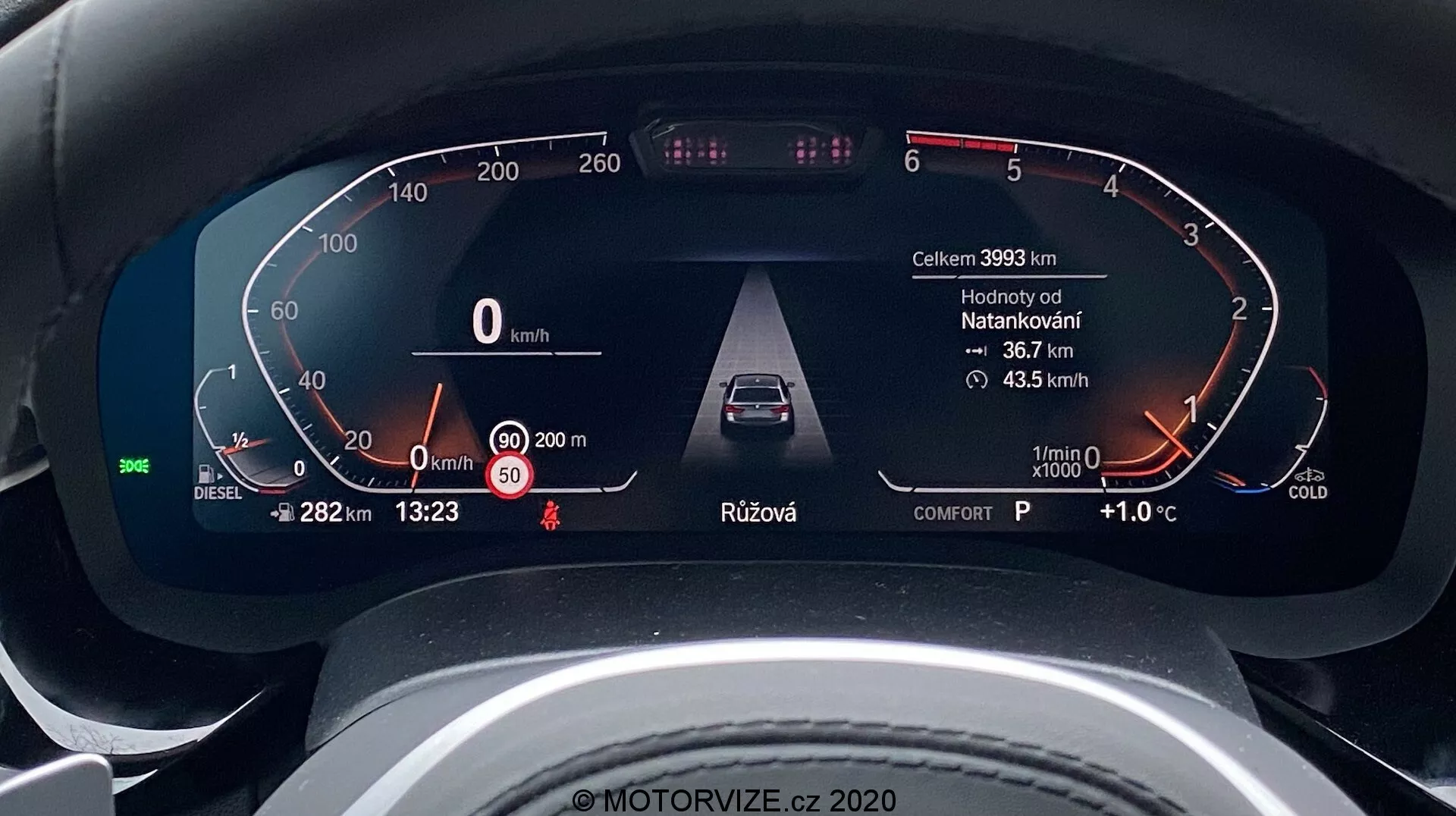 Közelkép egy 2020-as BMW 5-ös sorozat (BMW 520i Touring Steptronic) digitális műszerfaláról a vezető nézőpontjából. A digitális műszerkijelző kulcsfontosságú információkat jelenít meg, mint például a jármű sebessége (0 km/h), a dízelüzemanyagra utaló üzemanyagmérő, nyitott ajtó figyelmeztetés és biztonsági öv emlékeztető, fejlett vezetői segédrendszerek, mint az adaptív sebességtartó automatika, alapjárati fordulatszámmérő hideg motor jelzővel, valamint további adatok, mint a kilométerszámláló (39933 km), külső hőmérséklet (+1.0°C), idő (13:23), sebességfokozat választás (P parkoló pozíció) és vezetési mód (komfort). A dizájn elegáns és modern, ami egy felsőkategóriás járműre utal.