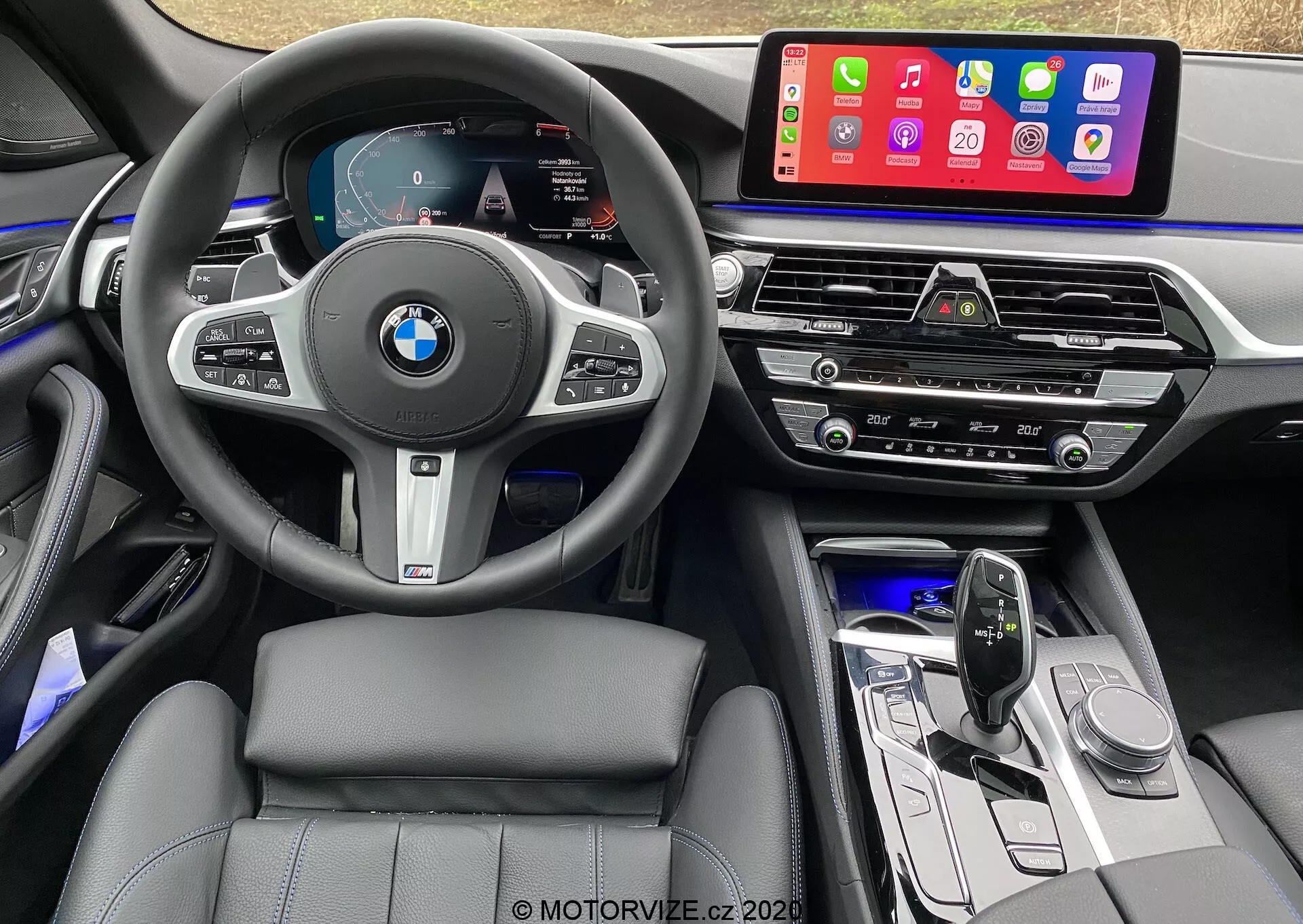 A 2020-as BMW 520i Touring Steptronic belső nézete a vezetőülésből, bőrbevonatú multifunkciós kormánykerékkel, BMW logóval és sebességtartó automatika, audió és telefonvezérlő gombokkal. A digitális műszercsoport sebességmérőt, fordulatszámmérőt és navigációs térképet jelenít meg. A középkonzolon egy nagy, érintőképernyős infotainment rendszer található, amely mobilalkalmazásokat, például az Apple CarPlayt jeleníti meg. Alattuk légbeömlők, klímavezérlő panel, audióvezérlők, elegáns kialakítású váltókar és funkciógombok találhatók. A belső térben bőr varrással ellátott ülések, hangulatvilágítás és magas minőségű, fémes díszítésű anyagok láthatók.