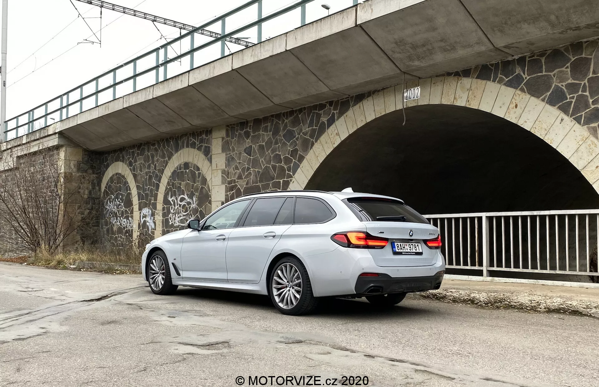 A képen egy modern, ezüst színű BMW 5-ös sorozatú Touring (520i Touring Steptronic) kombi látható, amely aszfaltos felületen parkol, háromnegyed hátsó nézetben. A jármű a kép jobb oldalán helyezkedik el, szinte párhuzamosan a kép alsó szélével. Az autó hátsó része, amely szembenéz a kamerával, megmutatja a hátsó lámpákat, a csomagtér ajtaját, a hátsó lökhárítót, valamint a jobb oldal nagy részét, beleértve a hátsó utas ajtaját, a jobb hátsó kereket és az első jobb ajtó és kerék egy részét. A jellegzetes dizájnelemek közé tartoznak a hátsó lámpák, a kettős kipufogórendszer, az ötágú könnyűfém kerekek, a nagy hátsó ablak és a tetőspoiler. A városi környezet többek között egy felüljárót és graffiti val díszített alagútbejáratot mutat kőműves munkákkal, borult ég alatt.