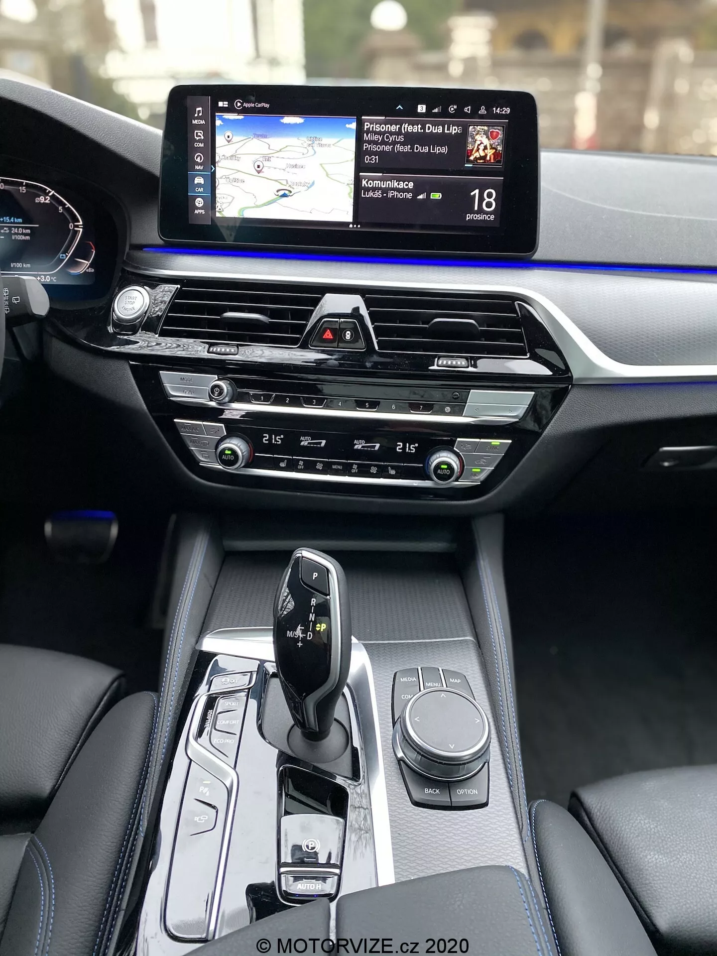 A BMW 5-ös sorozat 2020-as modell (BMW 520i Touring Steptronic) belső nézete, középkonzol és műszerfal a jobb első utas perspektívájából. A középkonzolon elegáns, automata sebességváltó váltókar 'park' üzemmódban, különböző vezérlőkkel, beleértve az infotainment rendszer forgótárcsáját és kapcsolókat a vezetési módok vagy járműbeállítások számára. A műszerfal középső része klímavezérlőkkel, intuitív gombokkal és tárcsákkal van ellátva, felette egy CD-nyílás található. A műszerfal felső részén egy széles infotainment érintőképernyő dominál, amelyen navigációs térkép és sebességhatár információk jelennek meg. Részben látható a digitális műszerfal, amely a sebességmérőt mutatja. A belső anyagok kiemelik a prémium minőséget, fekete bőr és kék díszítőcsíkok kontrasztja a műszerfal egész szélességében.