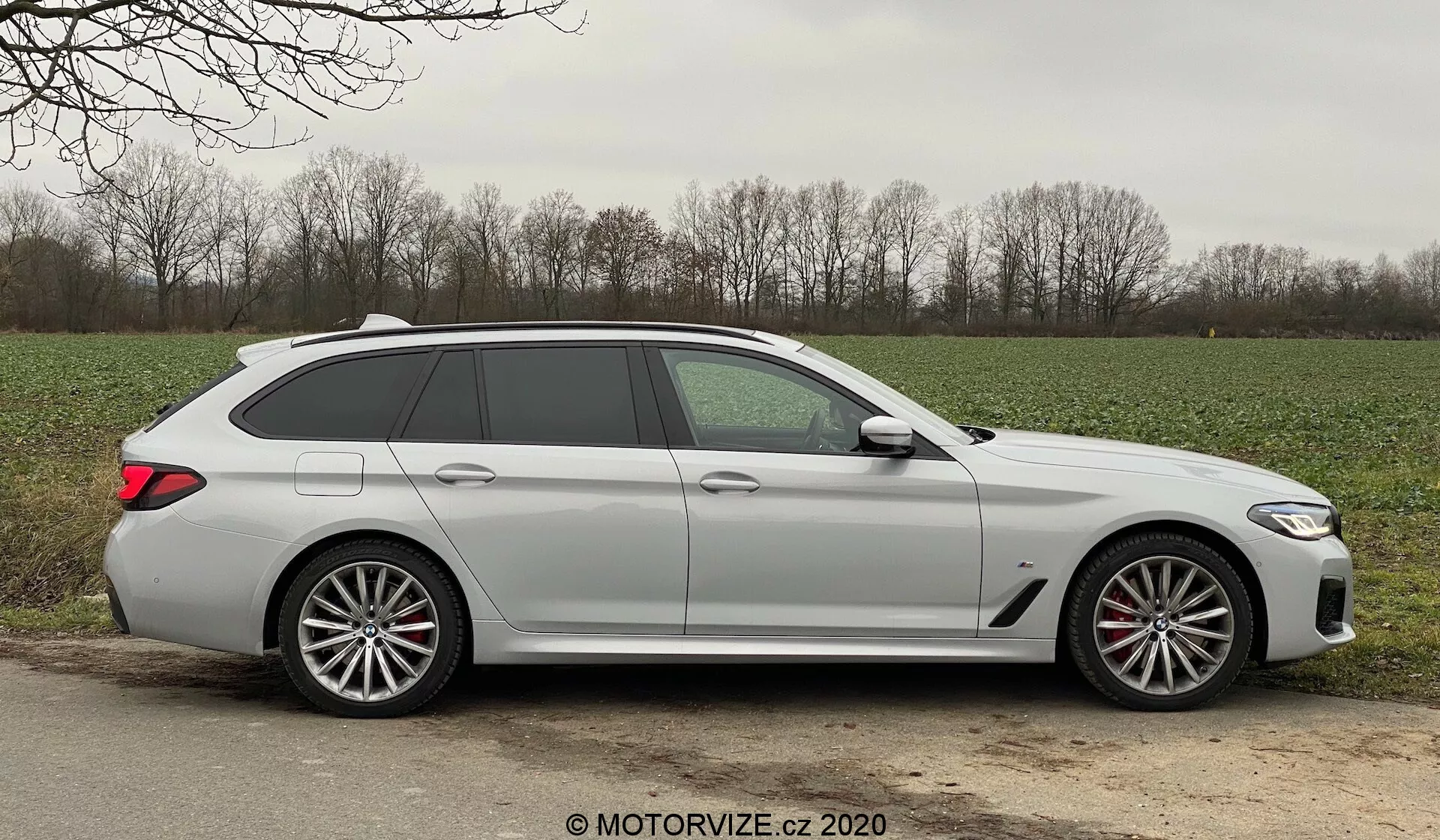 Egy modern BMW 520i Touring Steptronic kombi oldalnézete nappali fényben, amely elegáns dizájnvonalakat, öt ajtót, beleértve a hátsó ajtót is, hosszú tetővonalat tetőspoilerrel, nagy, többlazácsú könnyűfém kerekeket látható féknyergekkel, karosszéria színére fényezett ajtókilincseket, az oldalsó tükrökbe integrált irányjelzőket, enyhén sötétített ablakokat és matt vagy nem fémes festékfelületet mutat be. Az autó egy kavicsos vagy földes vállon van elhelyezve egy aszfaltozott út mellett, vidéki környezetben, fűvel és fákkal a háttérben, kedvező időjárási körülmények között.