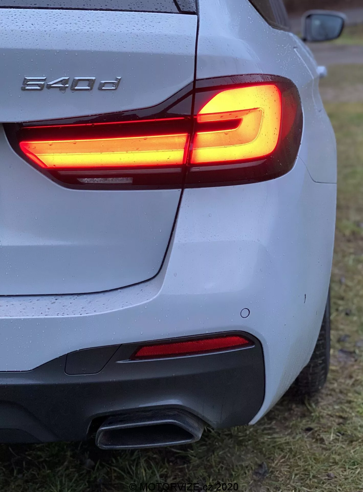 Közeli, háromb negyed hátsó szögből készült nézet egy BMW 540i-ről, amely bemutatja az L alakú LED-es hátsó lámpát, kettős kipufogócsöveket, ezüst/szürke karosszériát vízcseppekkel, formázott hátsó lökhárítót, részleges BMW jelvényt, füves területen parkolva.