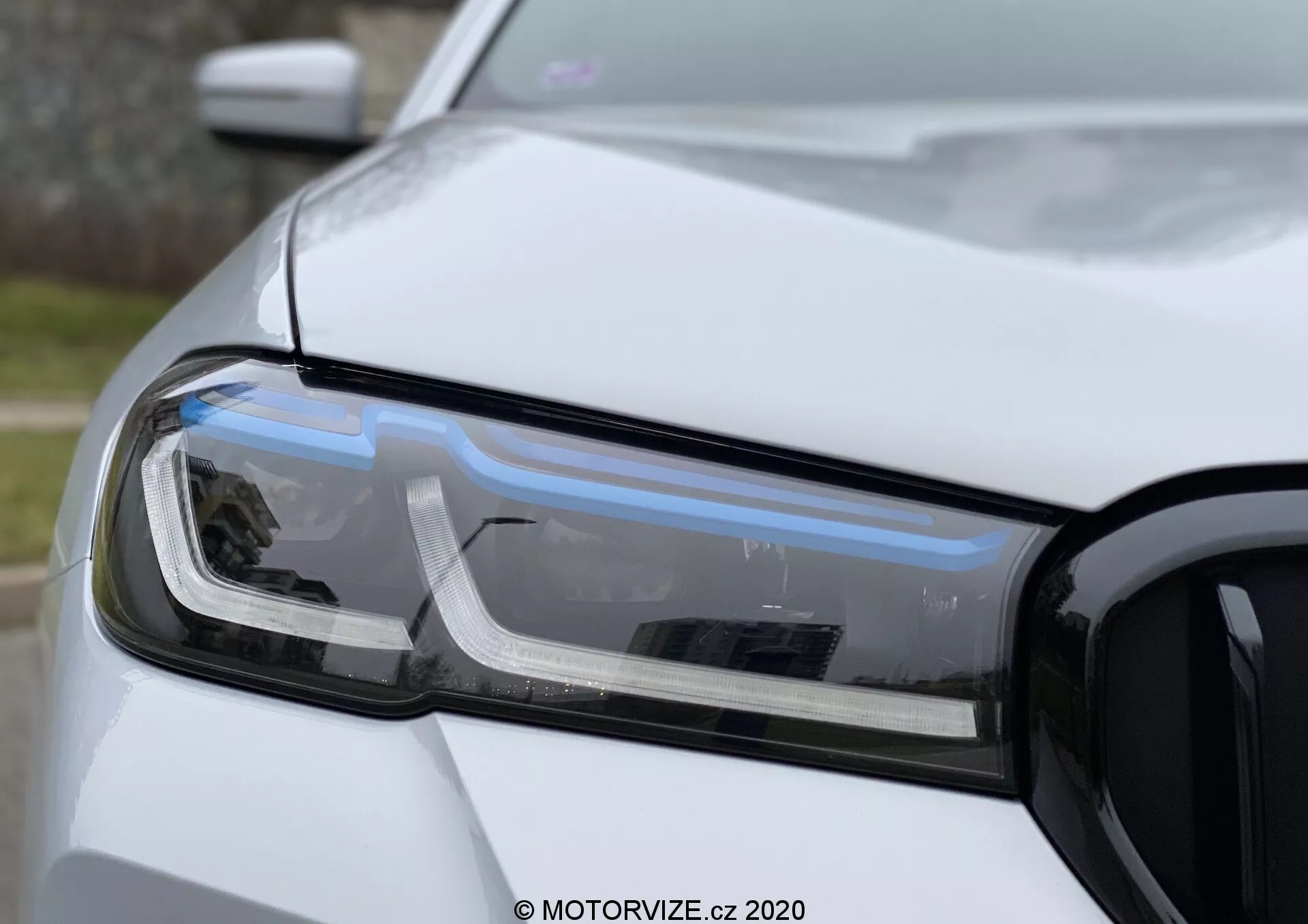 Közelkép a BMW 520i Touring Steptronic vezetőoldali fényszóró együtteséről, kiemelve a LED nappali menetfényeket, a projektoros fényszórót és az irányjelzőt. A fotón az autó bal első része látható, beleértve a motorháztető, az első lökhárító és a sárvédő egy részét, a jobb oldalon látható hűtőráccsal. A kép kis mélységélességet használ, hogy kiemelje a fényszóró technológiáját és dizájnját természetes, szórt fényben, szilárd, nem metál fehér festékbevonattal.