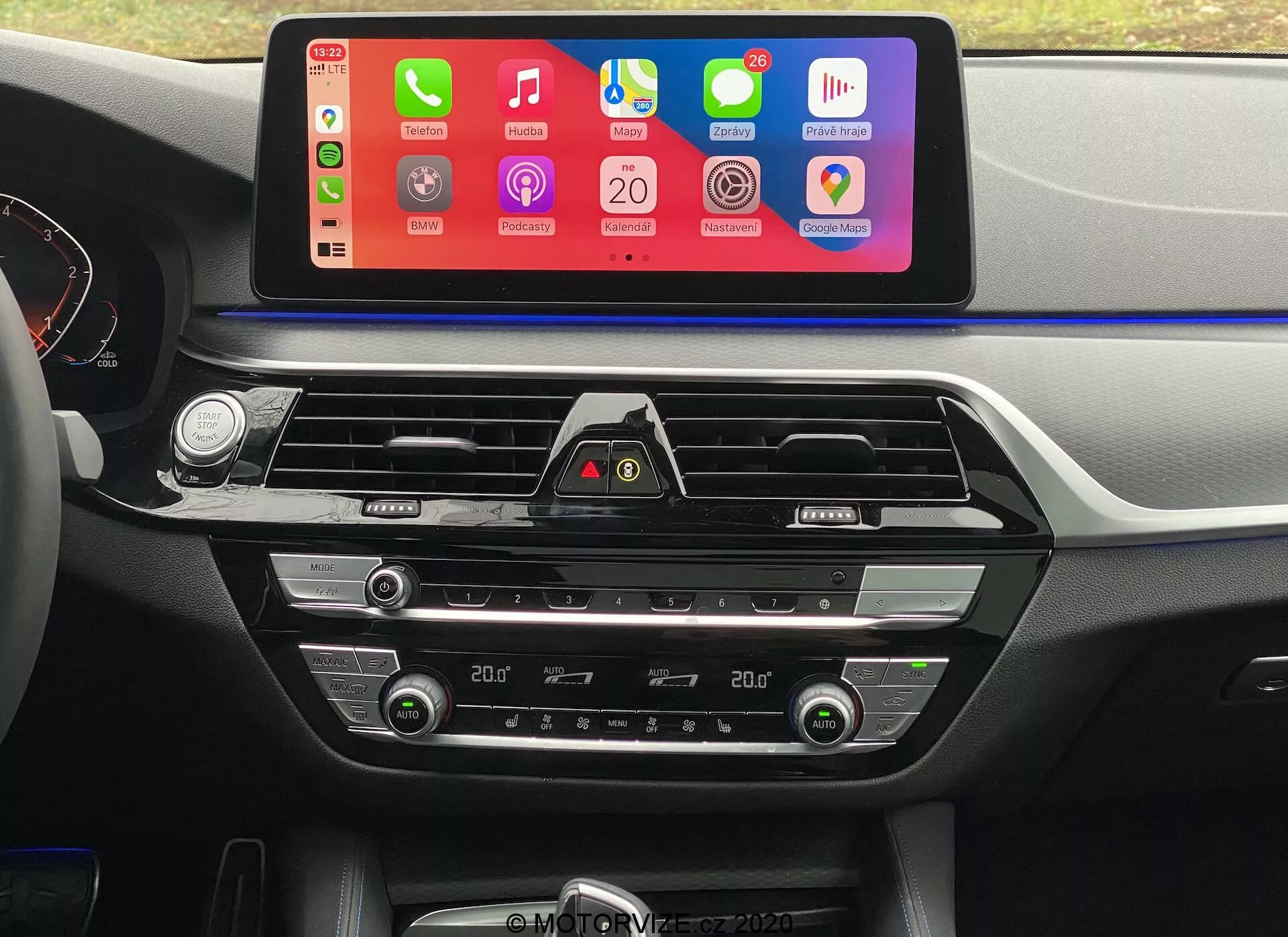 Egy BMW 5er-Reihe 2020 (BMW 520i Touring Steptronic) belső fotója a vezetőülés perspektívájából, a középső műszerfalra összpontosítva. A legkiemelkedőbb jellemző egy nagy, vízszintesen orientált infotainment érintőképernyő, amely színes felülettel rendelkezik, és támogatja a telefonintegrációt, mint például az Apple CarPlay és az Android Auto. Az érintőképernyő alatt krómozott szellőzők és egy vészvillogó gomb látható. Ezen kívül alul találhatók a kétzónás klímaberendezés vezérlői digitális kijelzőkkel a hőmérséklet beállításához, valamint gombok és tárcsák a levegő hőmérsékletének, ventilátor sebességének és levegőeloszlásának beállításához. A műszerfal és a középkonzol puha tapintású műanyagból és fémszerű díszítésekkel készültek, amelyek fokozzák az autó belső dizájnjának esztétikai vonzerejét.