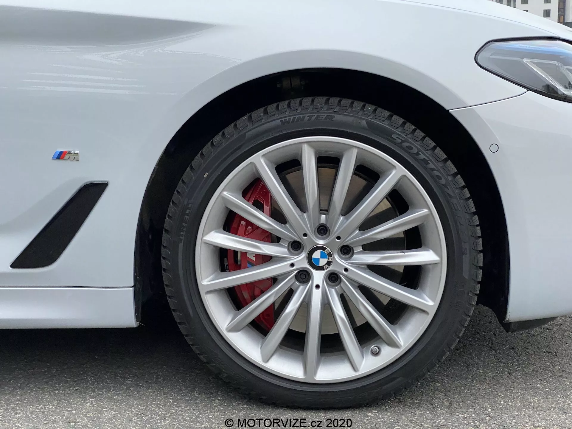 Közelkép egy 2020-as BMW 5-ös sorozat, konkrétan az 520i Touring Steptronic modelljének első kerék szakaszáról. Az alacsony szögből készült fénykép kiemeli a kerék összetett, többszörös küllőkkel ellátott ötvözött dizájnját ezüst kivitelben, amelyet egy piros féknyereg és egy szellőztetett rotor egészít ki. A BMW embléma és az 'M' jelvény az sárvédőn arra utal, hogy ez egy BMW M sorozat teljesítményorientált járműve. A kép kulcsfontosságú részleteket rögzít, amelyek alkalmasak nagy teljesítményű fékrendszerek, luxus jármű dizájnelemek és gumiabroncs-technológia bemutatására.
