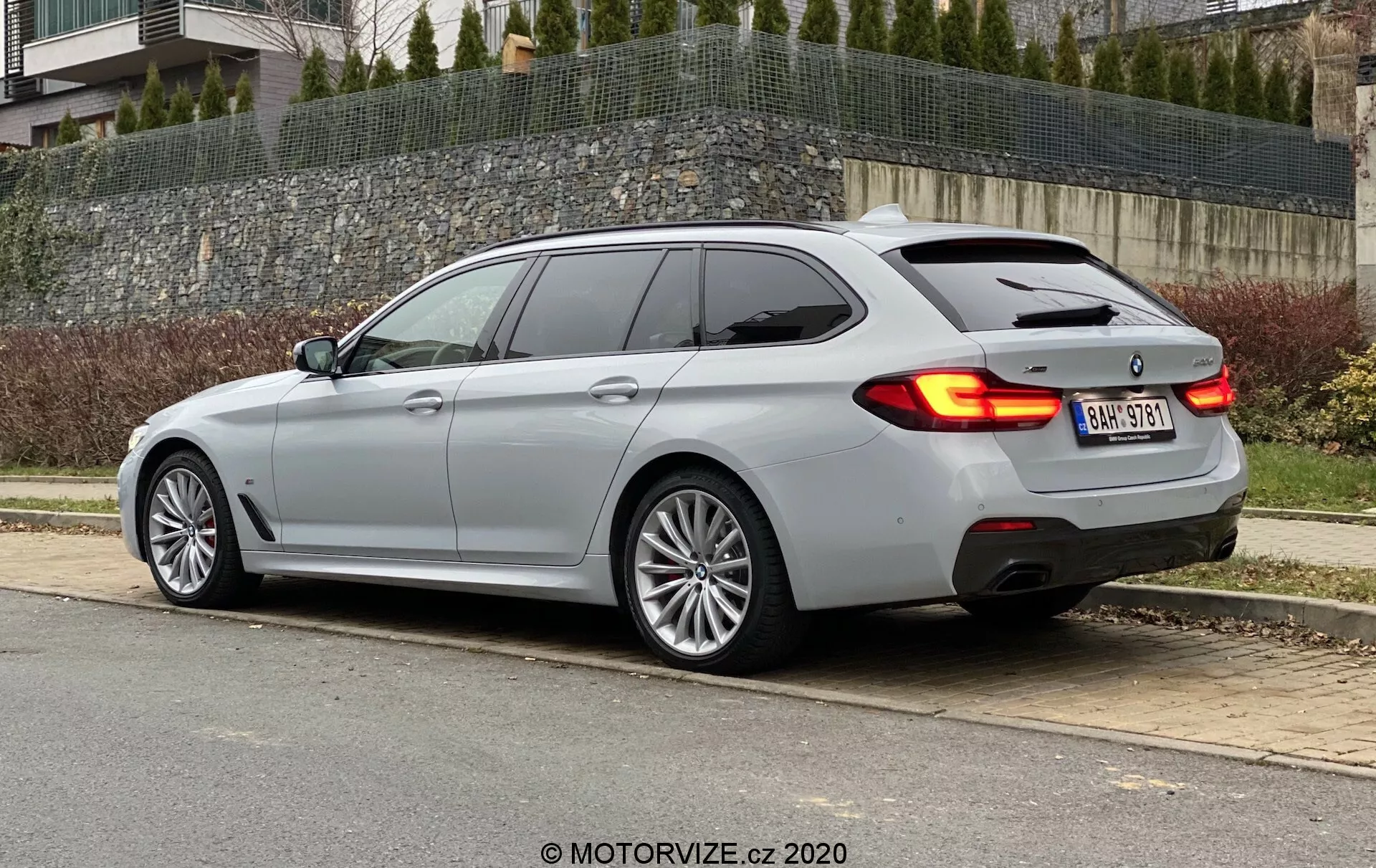 A képen egy modern kombi autó látható háromnegyed hátsó nézetből, amely a jármű elegáns tetővonalát és az oldalsó profilját mutatja. Kiemeli a vízszintes hátsó lámpákat, amelyek kissé belenyúlnak a csomagtérajtóba, és a meredeken lejtő hátsó ablakot, ami sportosabb megjelenést kölcsönöz, valamint króm díszítéseket az oldalsó ablakok körül. Az autó egy metálszürke felülettel rendelkezik, amely tükrözi a borult fényviszonyokat, többküllős kerekekkel utalva a luxus kategóriára, valamint kettős kipufogócsövekkel, ami erősebb motort vagy sportcsomagot sejtet. A LED hátsó lámpák és a finom hátsó légterelő jelenléte növeli a technológiai és sportos esztétikát.