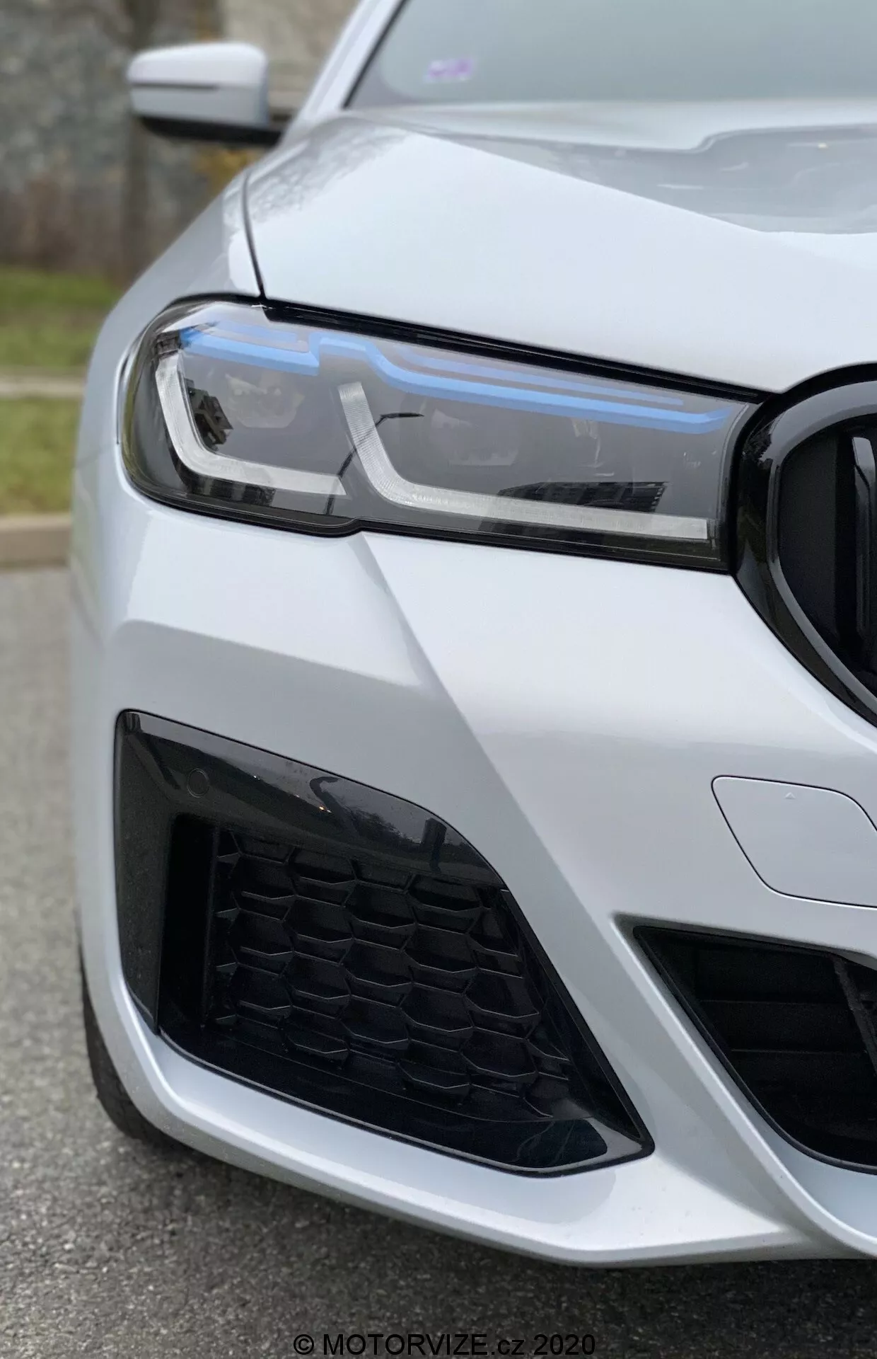 Közelkép egy BMW 520i Touring Steptronic (2020) autó bal elülső részéről, alacsony szögből fényképezve a fényszóróra, a hűtőrácsra és a lökhárítóra fókuszálva. A modern LED nappali menetfények, a méhsejt mintázatú hűtőrács és az agresszív légbeömlő dizájn sportos és fejlett járműre utalnak. Az autó fényes fehérre van festve, kiemelve annak luxus és jól karbantartott megjelenését.