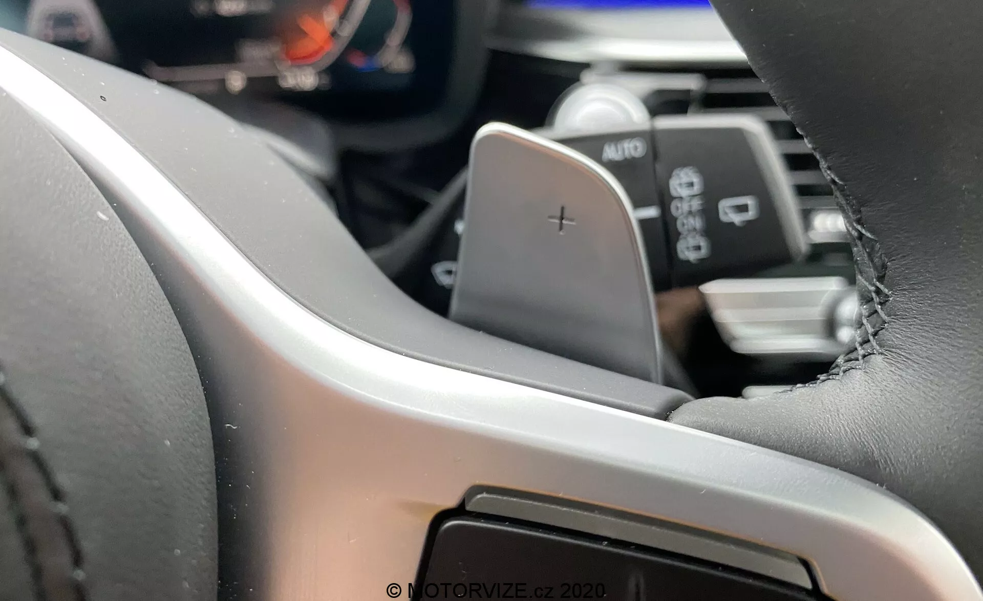 A BMW 5-ös sorozat 2020-as modelljének (BMW 520i Touring Steptronic) belső nézete, amely a kormánykerék bal oldalára összpontosít a vezetőülés perspektívájából⁠—modern, bőrrel bevont kormánykerék látható varrással, amely egy „+” jelű váltólapáttal van felszerelve. A digitális műszerfal vagy műszerpanel részben látható, kiemelve a fejlett vezetői információs rendszereket. A lapát alatt egy AUTO feliratú kar található, amely valószínűleg a fényszórók vagy az ablaktörlők vezérlőit jelzi. Az elmosódott háttérben az információs rendszer vagy a középkonzol részletei láthatók, ami érintőképernyős interfészre utal.