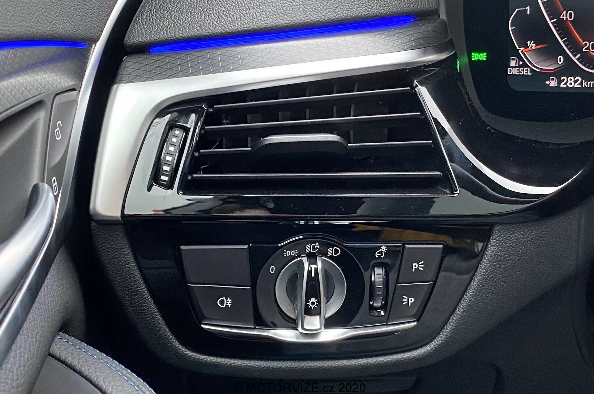 A 2020-as BMW 5-ös sorozat belső terének közeli képe, amely a vezetőoldali műszerfalra fókuszál. A vízszintes szellőzőnyílás felett kék hangulatvilágítás csík található. A központi vezérlők között van egy forgógomb az automatikus és tompított fények beállításához, gombok valószínűleg a ködlámpákhoz és a fényszórók szintezéséhez, további vezetőtámogató gombok és egy digitális sebességmérő, amely 0 km/h-t mutat parkoló módban lévő járművel és 282 km-es futásteljesítménnyel. Egy 'DIESEL' jelzés is látható.