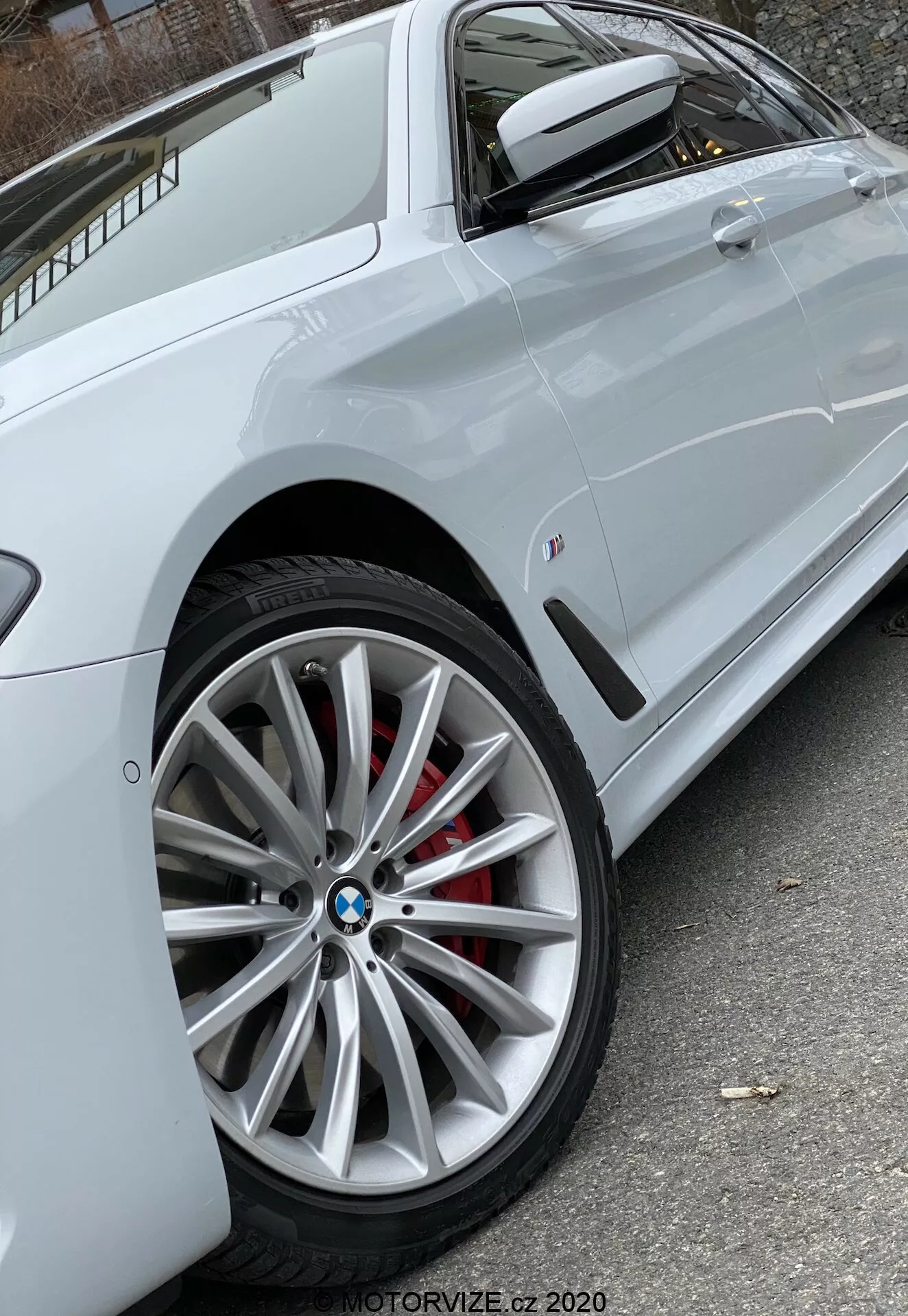 Közelről ferde oldalsó nézet egy fehér BMW 520i Touring Steptronic (2020) járműről, amely a modern többszálú ötvözetből készült első kereket emeli ki, látható BMW logóval és piros féknyeregkel, részben látható az oldalkaraktervonal, amely áthalad az ajtókilincseken, a jármű színével megegyező oldal tükörrel, integrált irányjelzővel, és egy kis plakett az első oldalpanelon. A természetes fény biztosítja a jármű jellemzőinek tiszta láthatóságát. Fotó a MOTORVIZE.cz 2020-tól.