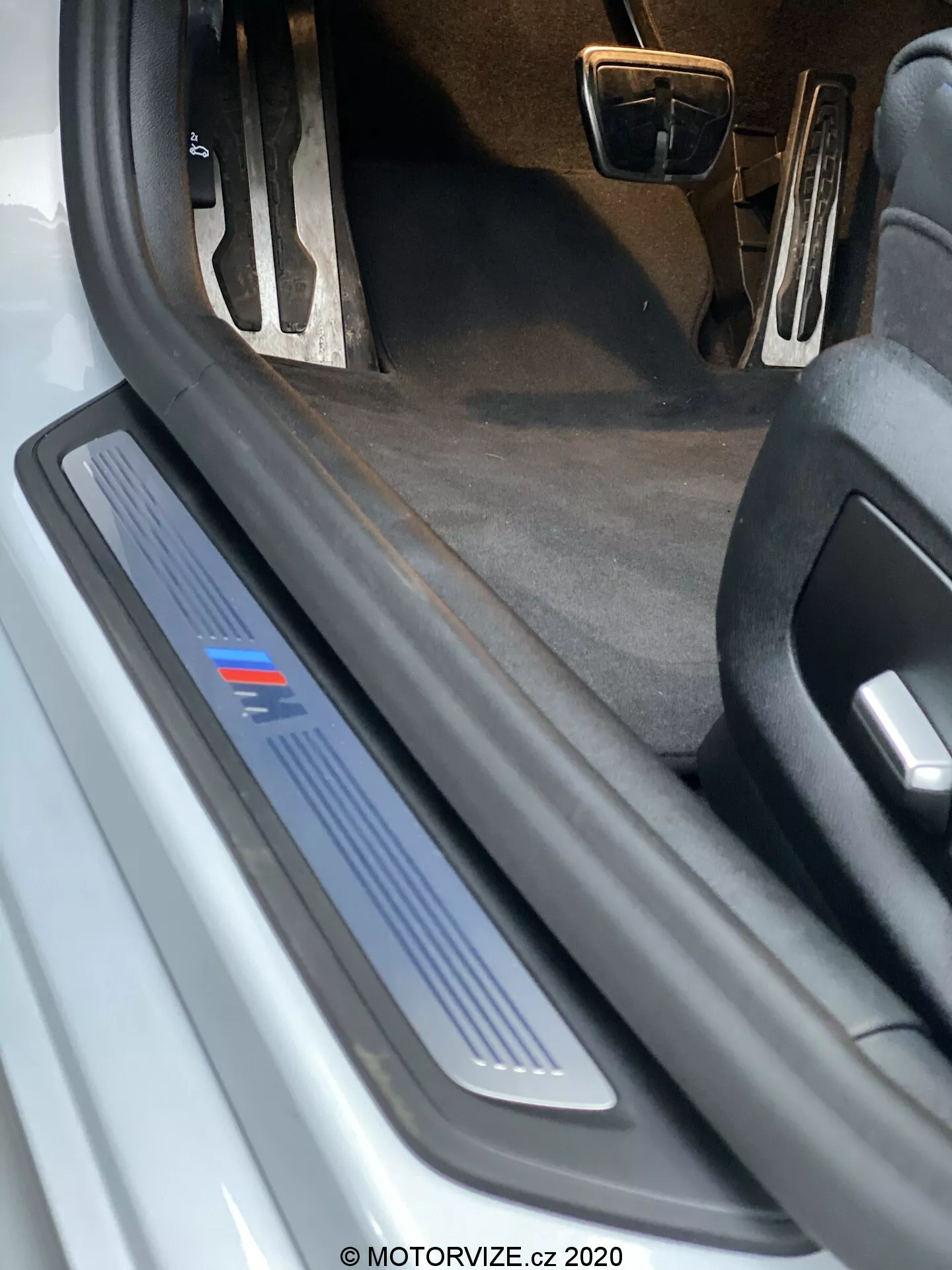 BMW 520i Touring Steptronic 2020 vezető oldalán lévő ajtóküszöb közeli nézete. A kép alacsony szögből készült, kiemelve a kék csíkkal díszített, kefélt fémből készült küszöbborítót, amely egy adott felszereltségi szintet vagy sportverziót jelezhet. A nyitott ajtó nem látható, de a védő és dekoratív küszöbborító nyilvánvaló, ami lehetséges LED világítást sejtet. A sötét szőnyeg belső és a részlegesen látható fémből készült pedálok sportos beltéri csomagot jelezhetnek. A küszöbborítón látható törmelékek és a kopás jelei arra utalnak, hogy az autó használt.