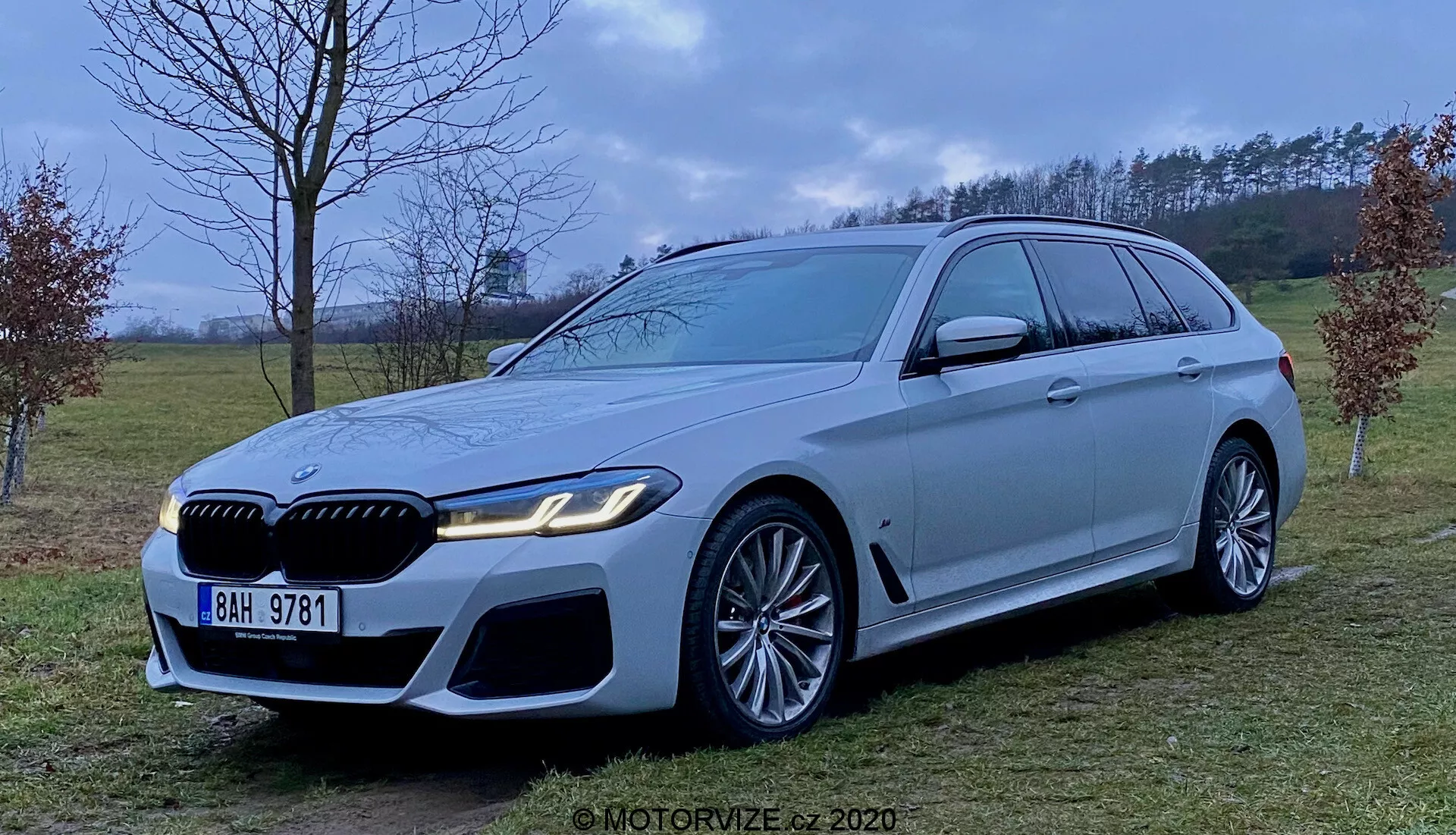 A 2020-as BMW 5-ös Touring (BMW 520i Touring Steptronic) luxuskombiról készült kép világos ezüst vagy szürke színű, agresszív és sportos első résszel, nagy légbeömlőkkel, kiemelkedő díszrácsokkal és elegáns LED fényszórókkal. A jármű nagy, sokküllős dizájnú könnyűfém keréktárcsákkal és jelentős féknyergekkel rendelkezik, hosszú tetővonallal és légterelővel, krómozott ablakkeretekkel és erős vállvonallal. A fénykép a jármű közepe magasságából készült, egy füves, félrurális környezetben, szórt fényviszonyok mellett.