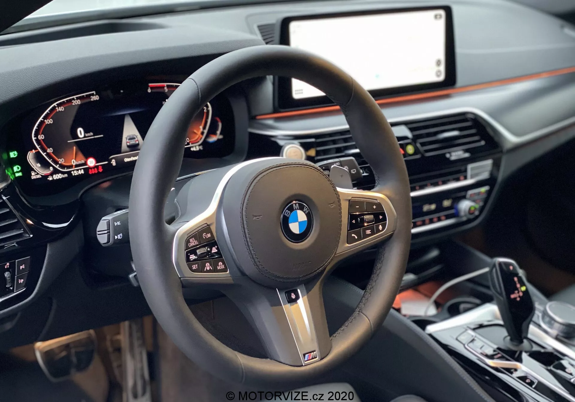 Első személyű nézet a vezetőülésből egy 2020-as BMW 520i Touring Steptronic autóban, a fókusz egy modern, háromküllős kormánykeréken van, vastag bőrbevonattal, multifunkciós gombokkal, valamint közepén egy kiemelkedő BMW logóval, amelyet fényes fekete díszítés és fémhatású elemek vesznek körül. A bal oldalon egy digitális műszeregység látható, amely sebességet, fordulatszámot és egyéb információkat jelenít meg. Jobb oldalon részben látható a központi infotainment képernyő. A sötét tónusú műszerfalon egy fa díszcsík is található, a képernyő alatti részen klímavezérlő és média rendszer gombokkal és tekerőkkel. A kép alsó részén a középkonzol látható, a sebességváltóval, iDrive vezérlővel és vezetési mód választó gombokkal.