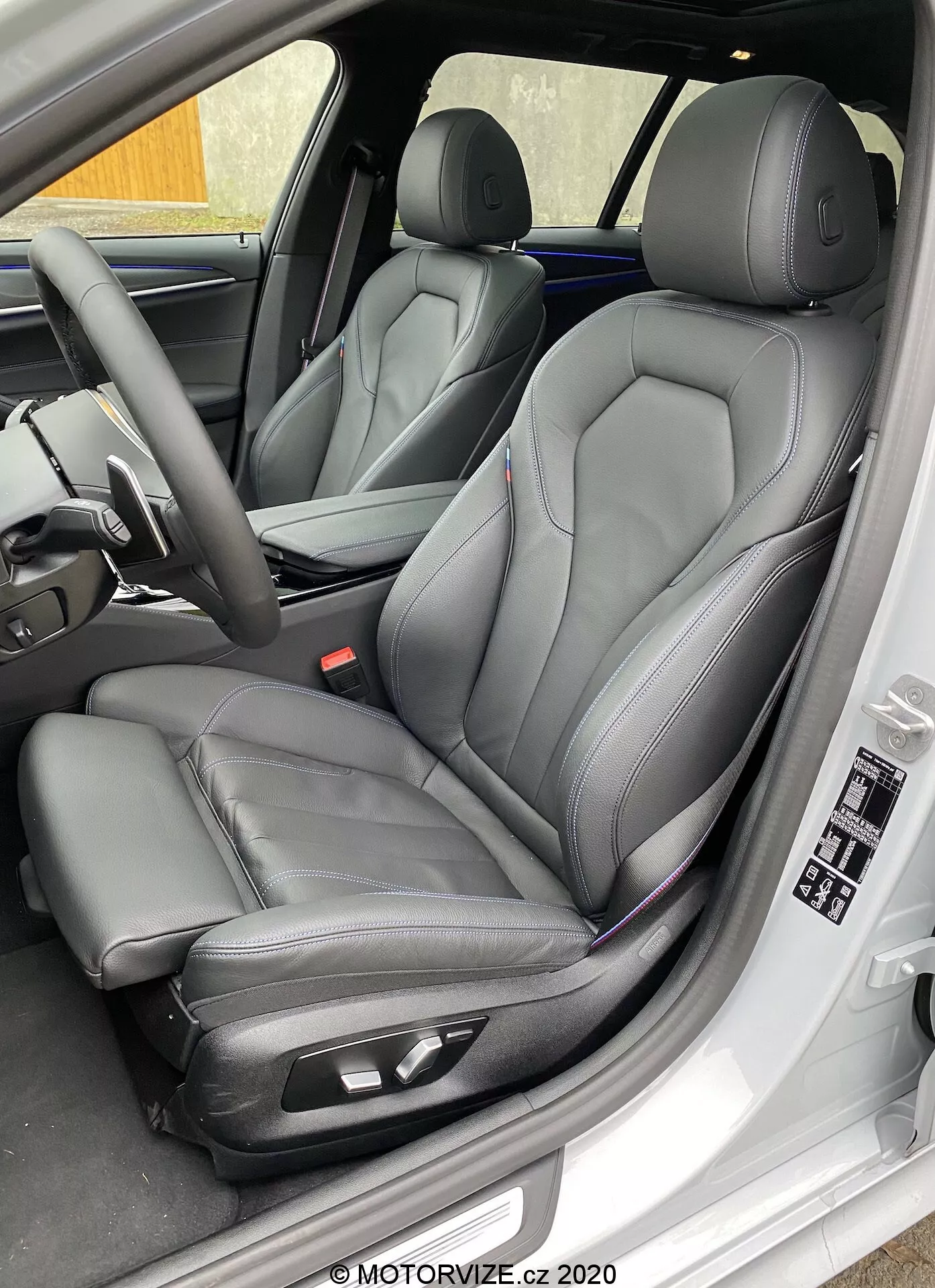 Közeli, enyhén szögletes oldalnézet a 2020-as BMW 520i Touring Steptronic vezetőüléséről, amely modern, fekete bőrrel és kontrasztos varrással készült kagylóüléseket mutat elektronikus állítási lehetőségekkel a kényelem érdekében. A képen látható továbbá a vezetői ajtó belső panelje elektromos ablak- és tükörvezérlőkkel, ülésmemória beállításokkal, valamint a műszerfal és a kormány további jellemzőivel, amelyek a luxust és a fejlett technológiát jelzik.