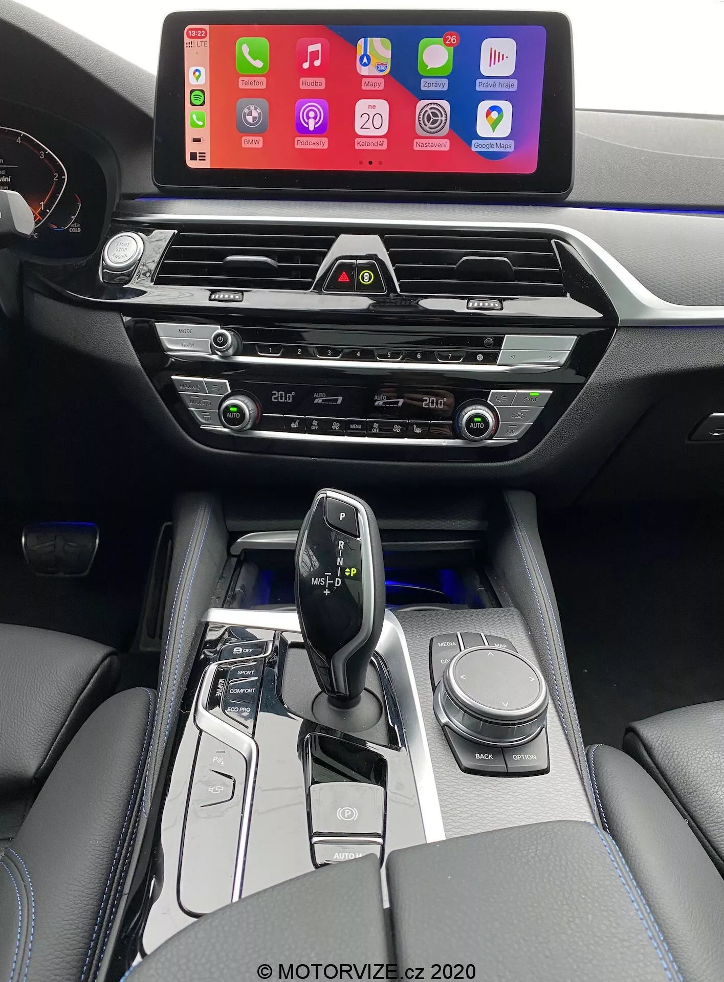 2020-as BMW 520i Touring belső fotója Steptronic váltóval, a vezető nézőpontjából kissé megemelve, hogy tisztább képet adjon a középkonzolról, a sebességváltó területéről és a műszerfal egy részéről. A kép egy nagy infotainment képernyőt mutat, amely okostelefon csatlakozási felületet jelenít meg, valószínűleg Apple CarPlay-t, magasan a műszerfalon, hogy könnyen látható legyen. Az infotainment képernyő alatt modern, vízszintes légbeömlők találhatók króm díszítéssel, alul pedig tapintható klímavezérlő gombok és tárcsák. A középkonzol tartalmazza az audiorendszer vezérlését, további klímavezérlőket és esetleg egy CD-lejátszó nyílást. Az ergonomikus és minimalista váltókar, amely valószínűleg elektronikus, magas fényű fekete és fém kiegészítőkkel van körülvéve, vezetési módválasztó gombokkal és egy navigációs forgótárcsával. Az autó belseje bőrkárpitozott karfákkal és látható öltésekkel rendelkezik, ami luxus érzetet ad. A váltókar körüli gombok lágy fehér megvilágítással kiemeltek, ami automata váltót jelez.