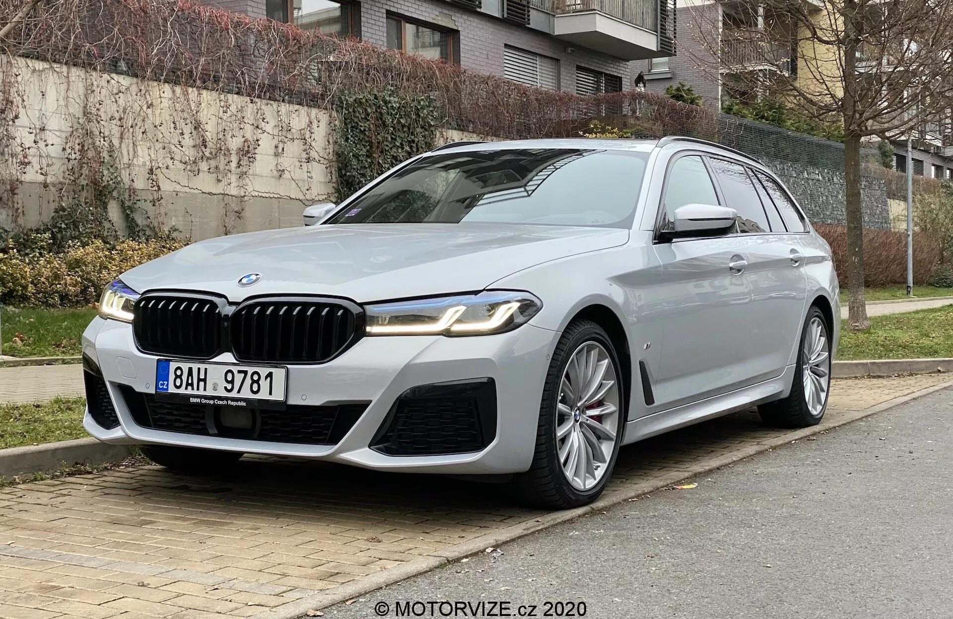 Luxus BMW 520i Touring parkol a városi utcán, alacsony elülső háromnegyedes szögből nézve kiemelve az autó elejét és a vezető oldalát. Az autót jellegzetes veserács jellemzi króm részletekkel, karcsú LED fényszórók, sportos első lökhárító és határozott motorháztető ívek. Többrudas könnyűfém kerekek, irányjelzőkkel és kamerával ellátott oldalsó tükrök, króm ablakkeretek és napfénytető adják a luxus megjelenést. A jobb oldalon elhelyezve, fűvel, bokrokkal és lakóépületekkel a háttérben, ami fejlett technológiát és magas szintű dizájnt sugall.