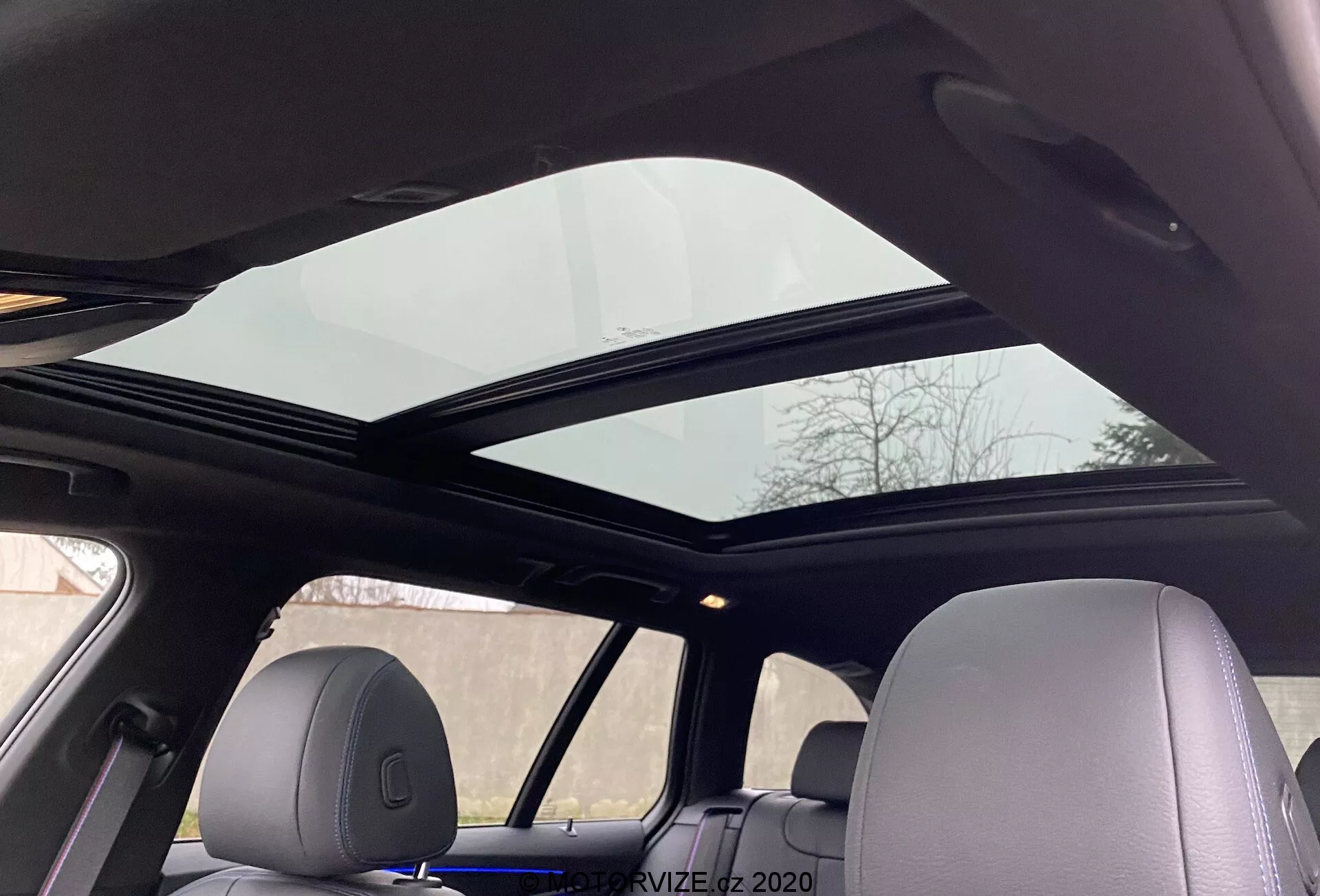 2020-as BMW 5-ös sorozat 520i Touring Steptronic váltóval, panorámás napfénytetővel a belső térben, az első és hátsó ülések fölött a hátsó ülésen ülő személy szemszögéből nézve. A napfénytető elülső panelje kissé megdöntve nyitva van, javítva a szellőzést. A fejliner világos színű, kiegészítve a jármű következetes belső kialakítását, az anyaghoz illeszkedő fejtámlákkal, hangsúlyozva a fejlett kényelmet és a modern kényelmi funkciókat.