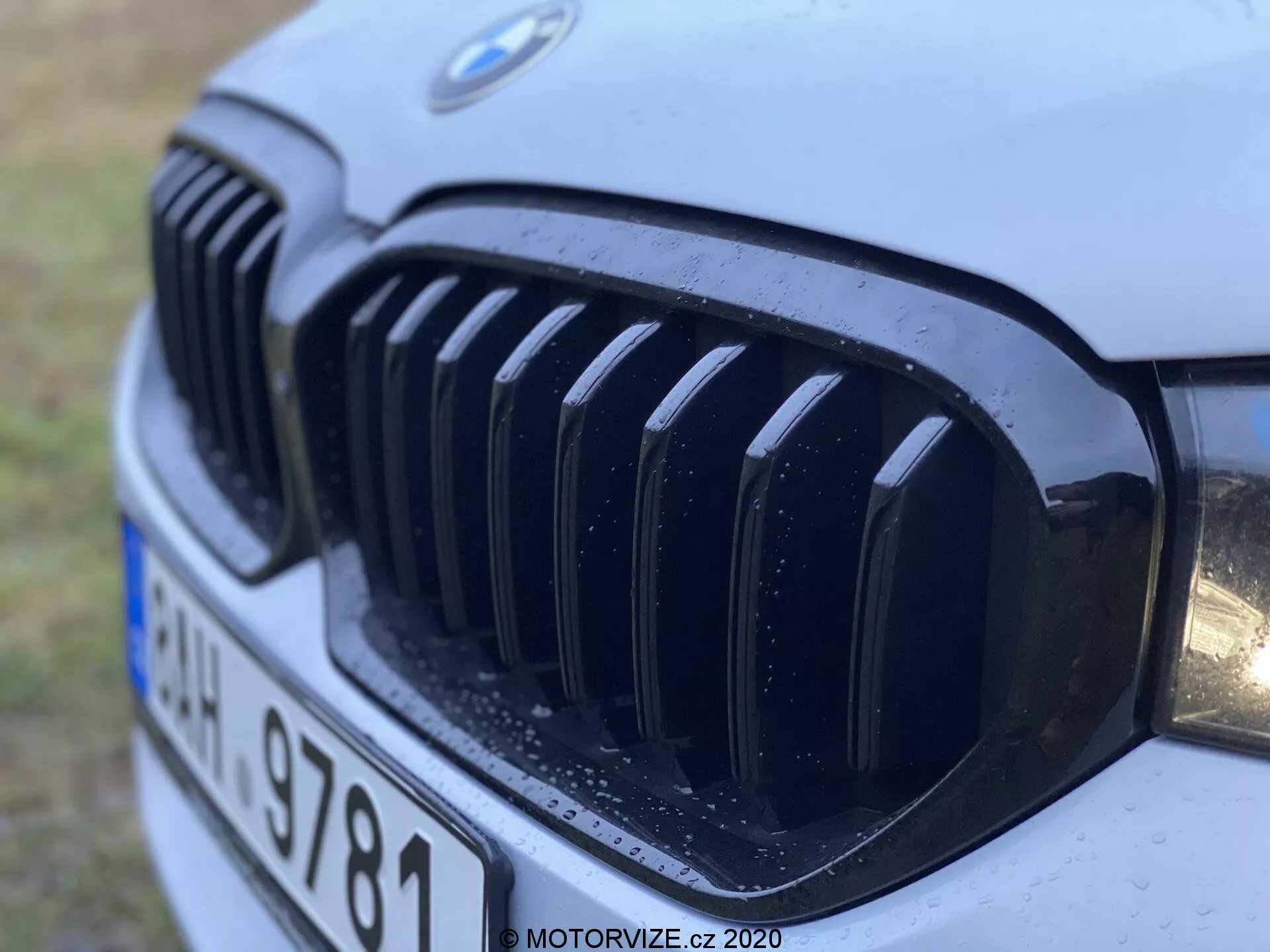 A 2020-as évjáratú BMW 520i Touring Steptronic motor hátsó rácsának közeli képe, amely bemutatja a jellegzetes veserácsot és a BMW emblémáját fekete függőleges lécekkel, részleges fényszóróval és vízcseppekkel, amelyek az eső vagy a mosás közelmúltbeli következményeire utalnak, alacsony, elülső-bal szögben készítve.