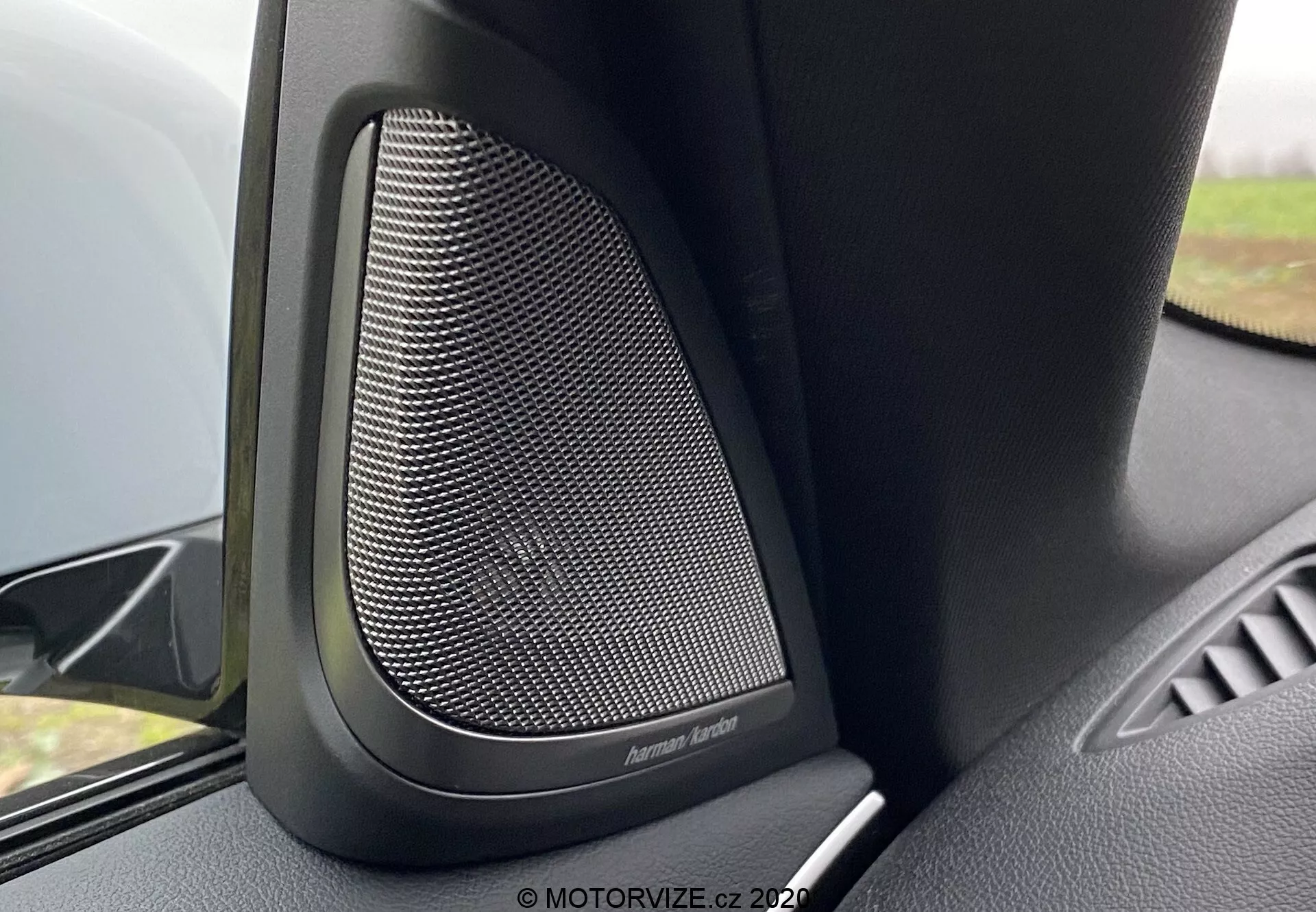 Közelkép a 2020-as BMW 5er-Reihe (BMW 520i Touring Steptronic) belső teréről, különös tekintettel a Harman Kardon prémium ajtóhangszóróra. A kép megörökíti a fém hangszórórács finom hálómintáját, amelyet fekete műanyag vagy puha anyagok vesznek körül a nem kívánt rezgések csökkentése érdekében, javítva a hangminőséget. A kamera szöge a felső ajtópanelt mutatja kiemelt hangszóróval és az ajtó kartámaszának egy kis részletével. A lágy háttérelmosás kiemeli ezt a high-end audiorendszert ebben a luxus járműben.