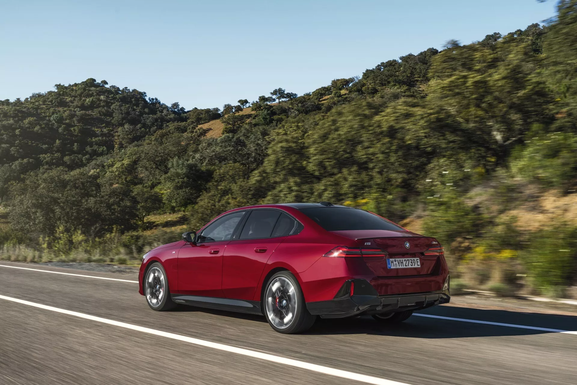 Piros, négyajtós BMW 5-ös sorozat (G60-G61) sedan mozgásban, háromnegyedes hátsó szögből rögzítve, kiemelve a hátsó és oldalsó design elemeket, mint a csomagtartó forma, hátsó lökhárító, LED hátsó lámpák és aerodinamikai vonalak egy hegyvidéki háttérrel tiszta égbolt alatt