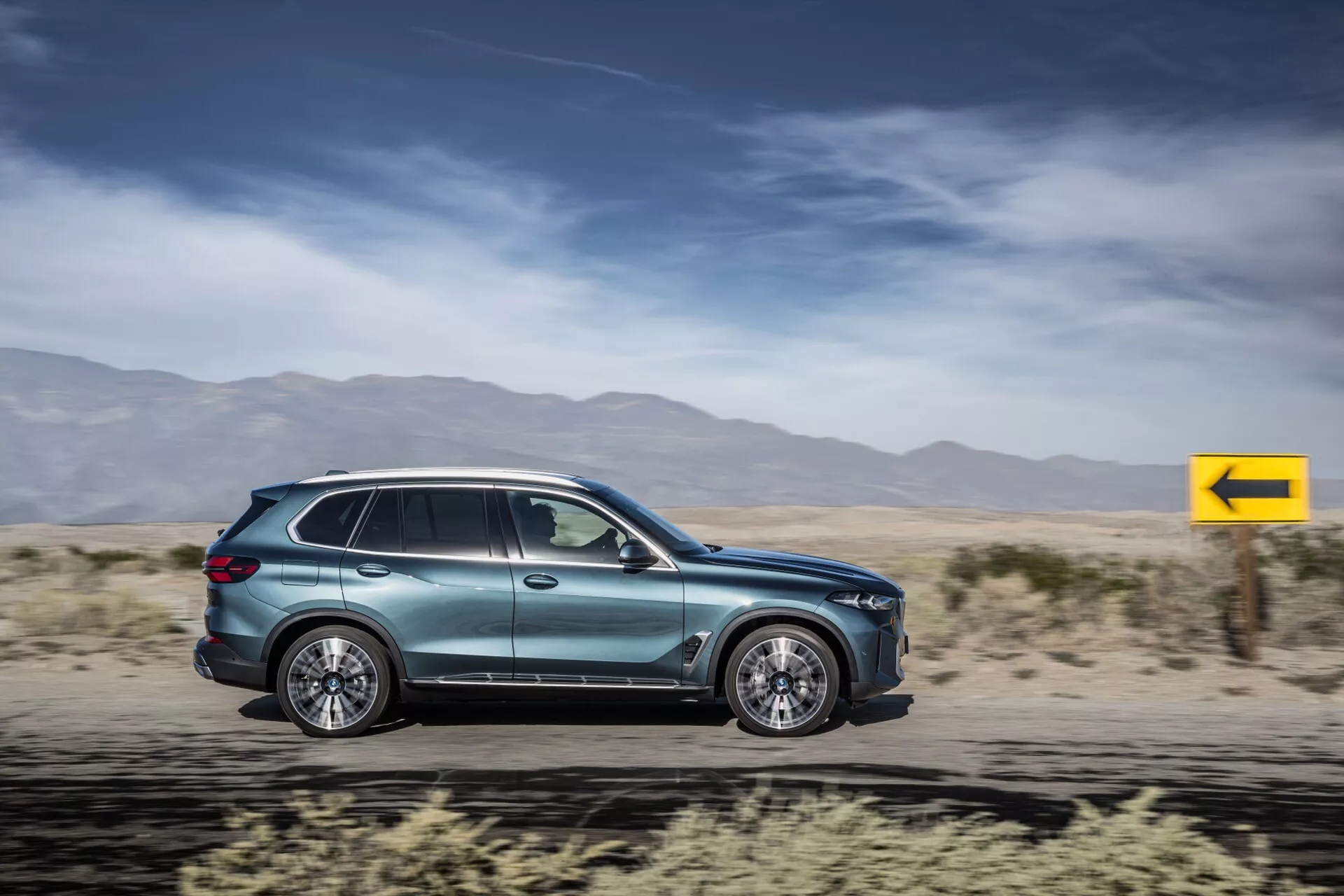 2023-as BMW X5 (G05-F95-Facelift) SUV mozgásban, bemutatva az aerodinamikát, többsugaras ötvözött kerekek, integrált ajtókilincsek, aerodinamikus oldalsó tükrök, sötétített ablakok, oldalsó jelző, fémes fényezés és modern hátsó lámpák dizájnt elmosódott sivatagi háttérrel szemben.