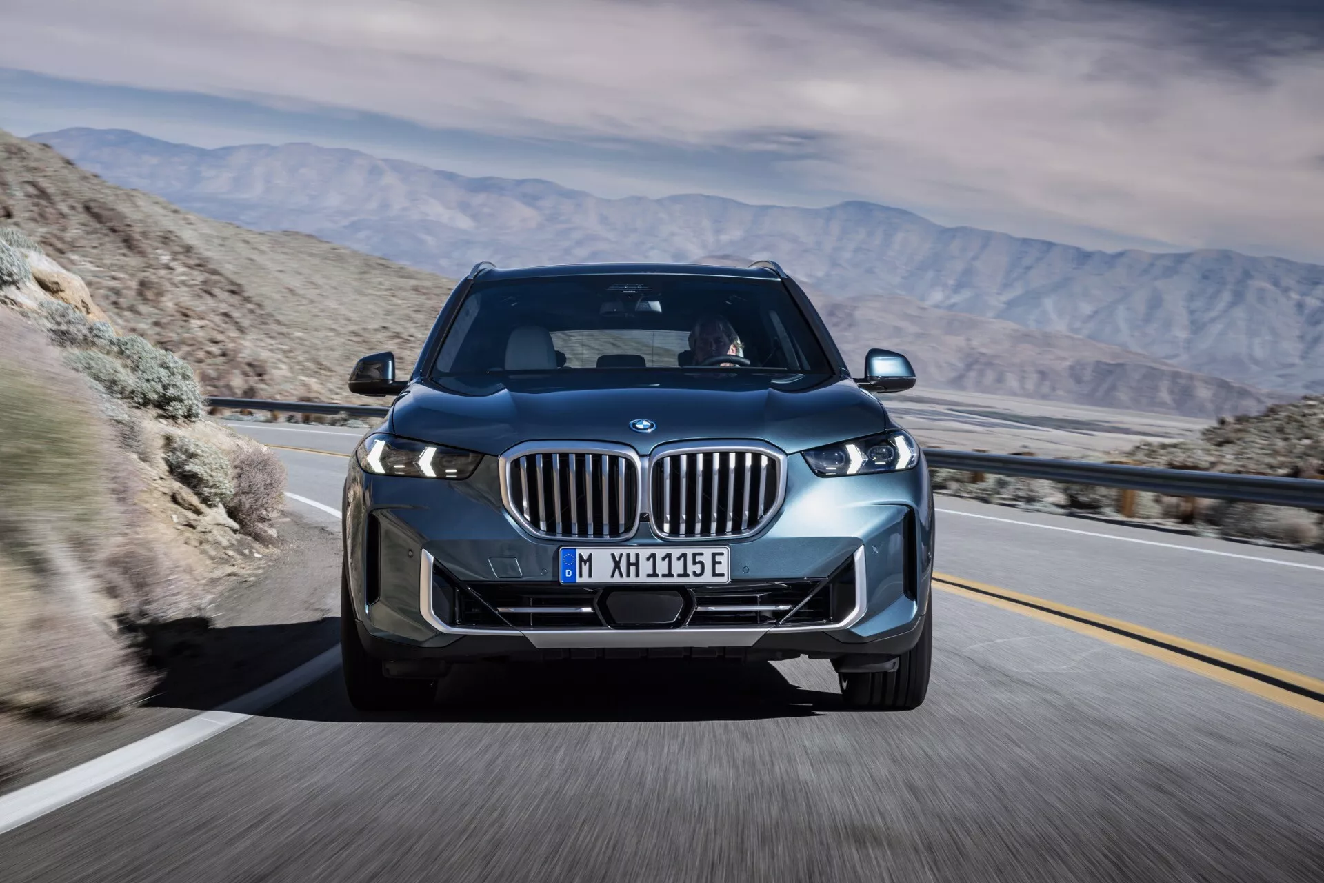 2023-as BMW X5 (G05 F95 modellfrissítés) mozgásban, háromnegyedes előlnézetből fényképezve, kiemelve az LED fényszóróit, vesemedence alakú rácsát aktív zsalukkal, ötvözött kerekeit és a sík aerodinamikai kialakítását egy hegyes sivatagi háttérrel szemben, amely fejlett technológiát és potenciális elektromos vagy hibrid képességeket sugall