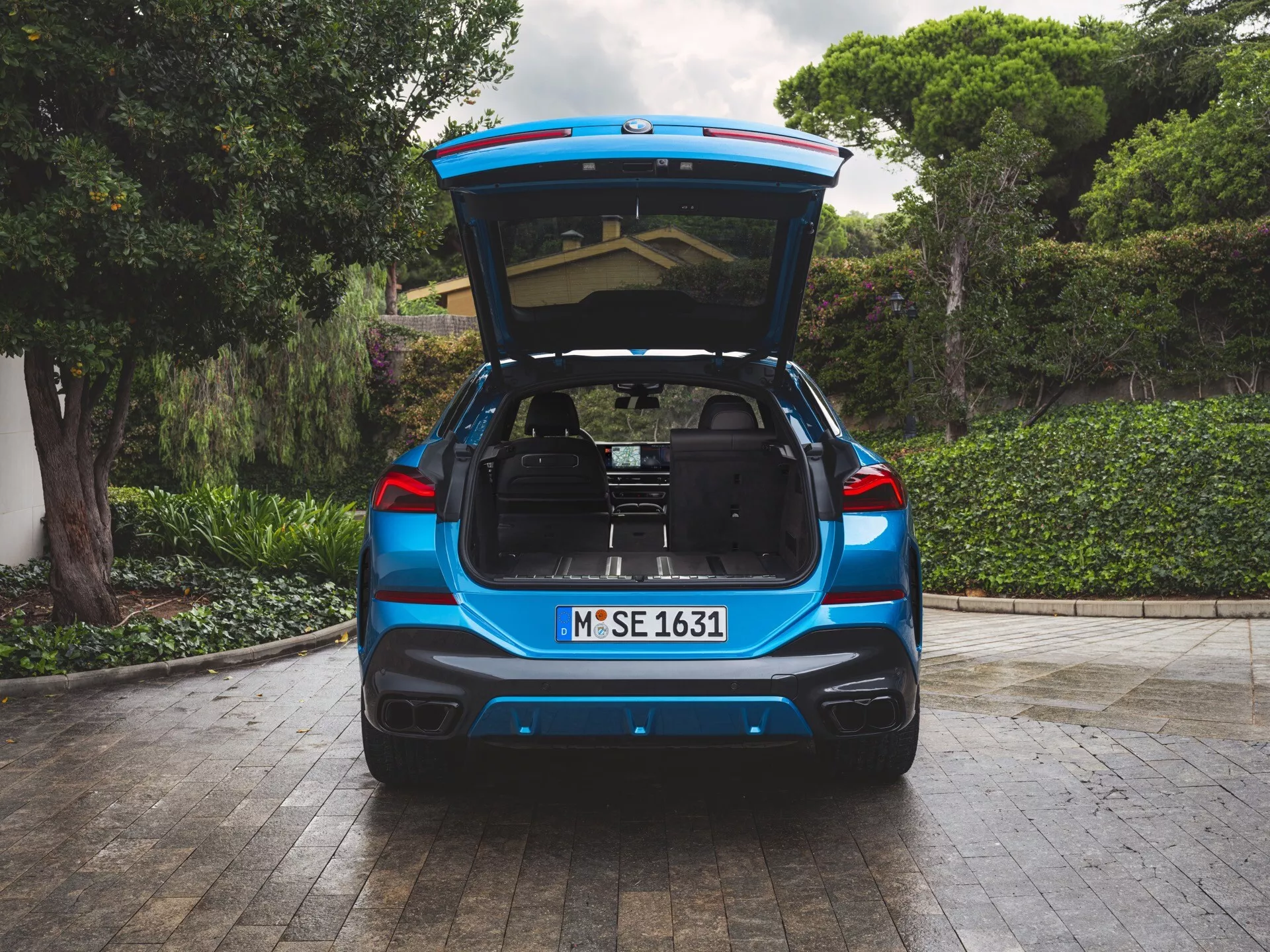 A 2023-as BMW X6 (G06-F96-facelift) hátsó nézete, kék metál fényezéssel, nyitott csomagtartóval, amely tágas rakodóteret tár fel, kőburkolatú területen parkolva, zöld növényzettel és egy ház részével a háttérben, LED hátsó lámpákkal, kettős kipufogókkal és lehetséges parkolást segítő technológiákkal