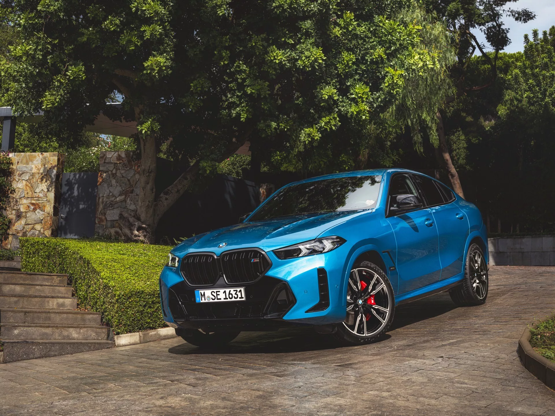2023-as BMW X6 (G06-F96-Facelift) kék színben, szögben parkolva, bemutatva előlapját, vese alakú hűtőrácsát, LED vagy lézer fényszóróit, sportos ötvözött kerekeit piros féknyergekkel, kontúros motorháztető vonalait és aerodinamikai oldalsó tükröket integrált irányjelzőkkel, természetes nappali fényben rögzítve zöld növényzettel és építészeti lépcsőkkel a háttérben
