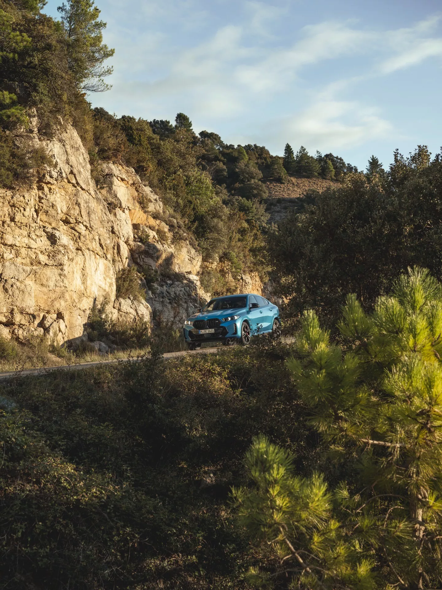 2023-as BMW X6 (G06-F96 modellfrissítés) háromnegyedes elölnézetben, egy kanyargós hegyi úton, erdővel körülvéve, bemutatva a terepjáró képességét és modern dizájnját