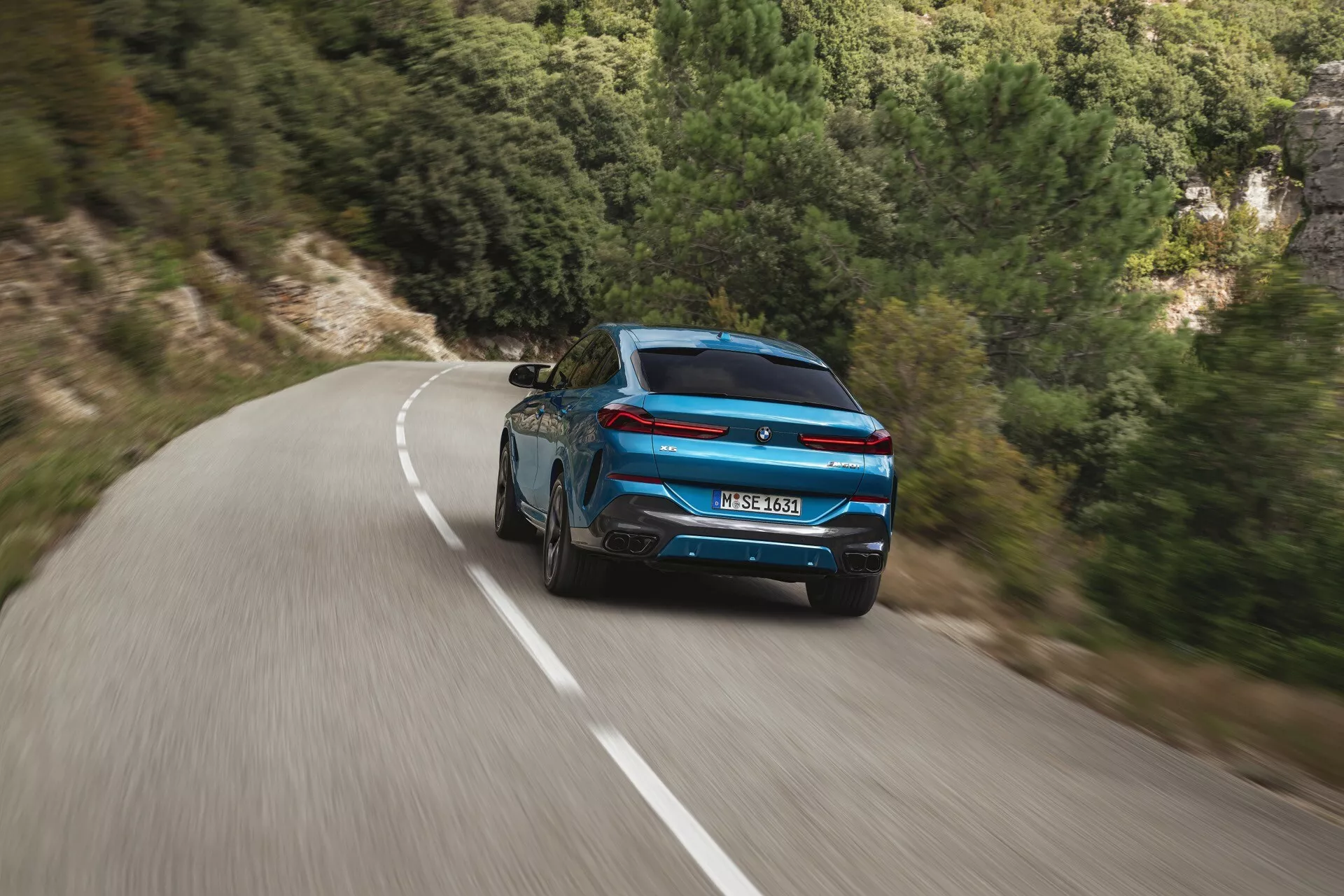 Dinamikus hátsó nézet egy kék BMW X6 2023 (G06-F96-Facelift) sportos kialakítással és modern hátsó lámpákkal egy kanyargós úton természetes tájkép közepette