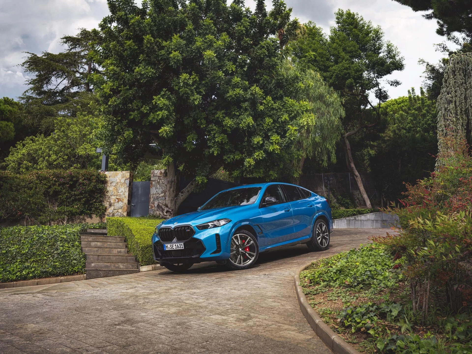 2023-as BMW X6 (G06-F96-Modellfrissítés) kék színben, háromnegyedes elölnézetben, egy beállóval körülvett zöld környezetben parkolva, bemutatva az első lökhárítót, hűtőrácsot, motorháztetőt, szélvédőt, a vezető oldali profilt, LED fényszórókat, sportos kerekeket piros féknyergekkel, és aerodinamikai kialakítást