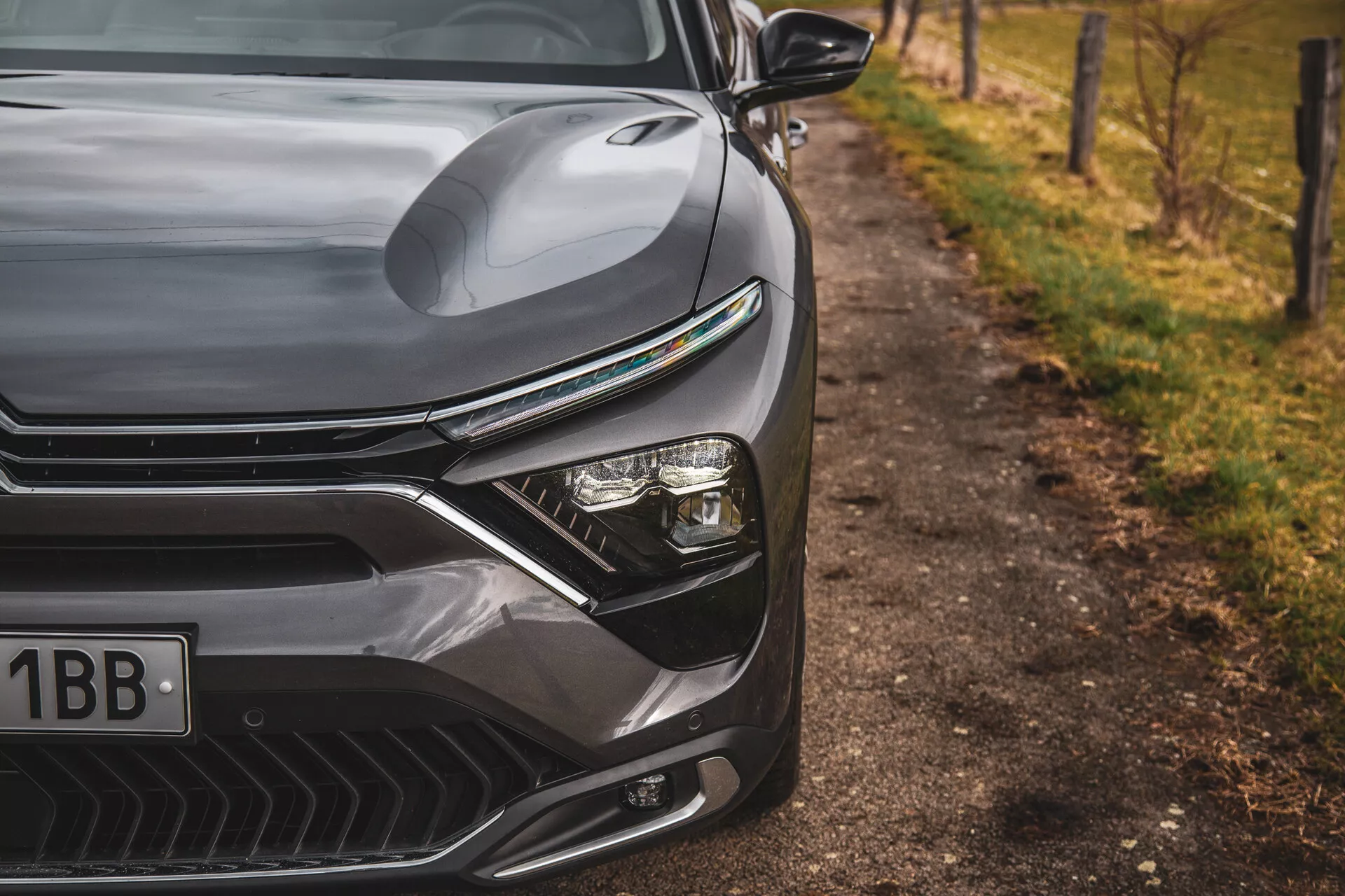 Közeli felvétel a 2022-es Citroen C5-X-ről, amely bemutatja az elegáns dizájnt az első bal oldalon, beleértve a kontúros motorháztetőt, a markáns hűtőrácsot vízszintes vonalakkal, a fejlett LED fényszóró rendszert, és a légbeömlőkkel ellátott aerodinamikus első lökhárítót, természetes útmenti háttérrel homályosítva.