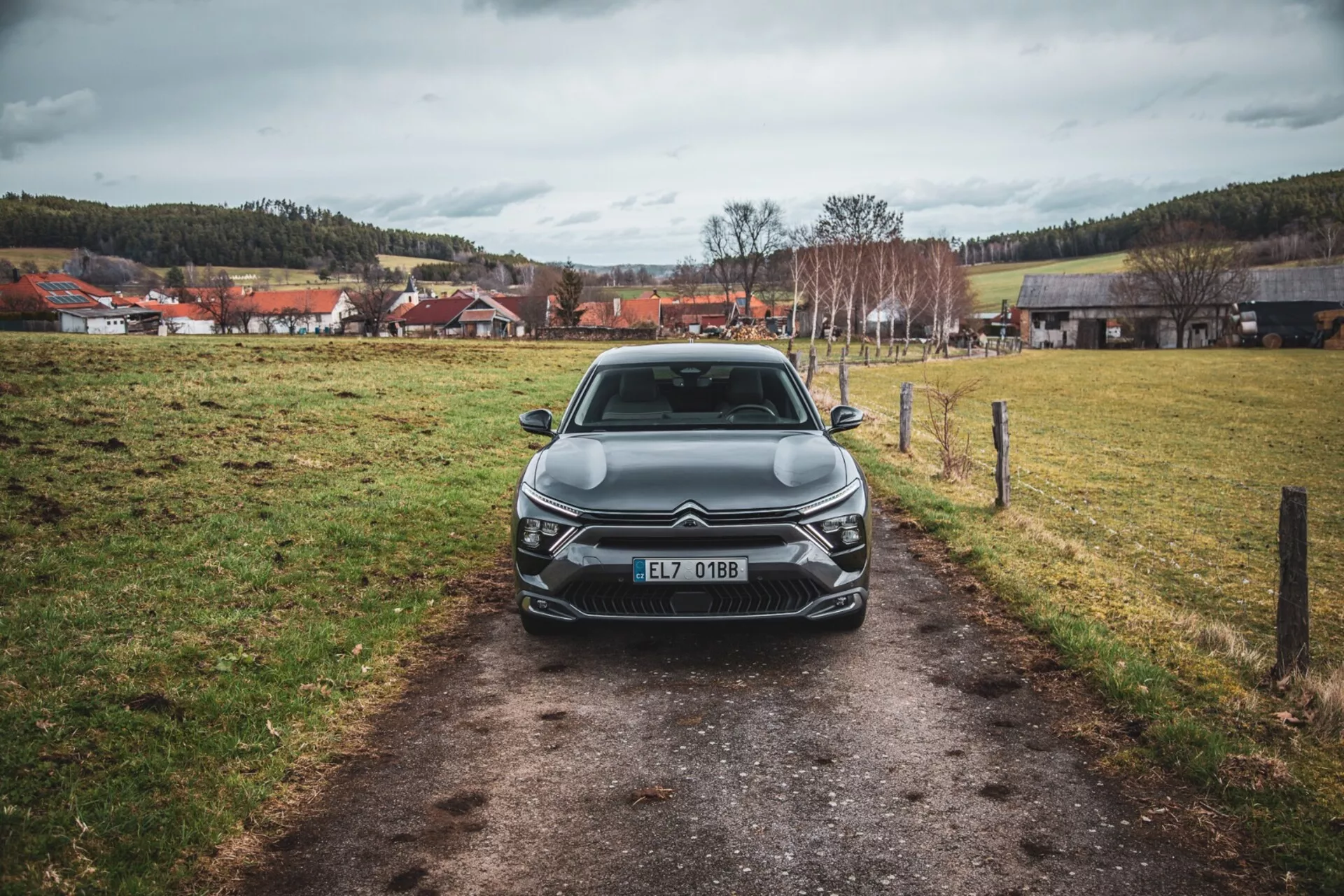 Egy 2022-es Citroën C5-X frontális nézete egy keskeny ösvényen, füves mezőn, borult égbolt alatt, bemutatva annak elegáns tervezését, bonyolult hűtőrácsát az emblémával, szögletes LED-es fényszóró egységeit, szobrászati vonalait a motorháztetőn, és további első világító elemeket, Cseh Köztársaságban regisztrálva.