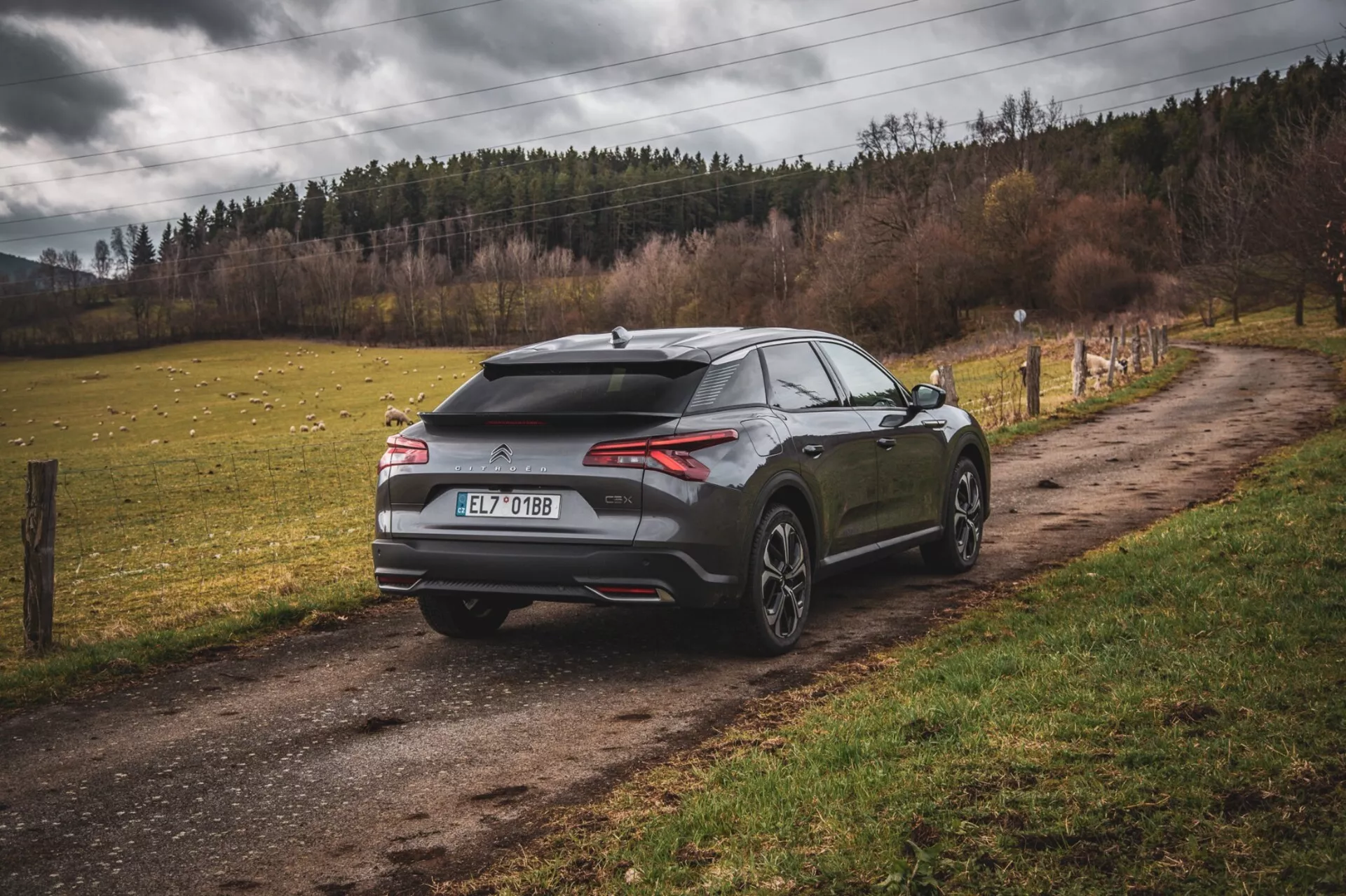 2022-es Citroen C5-X modern ferdehátú parkol egy murvás vidéki úton, hátulról és oldalról nézve, hangsúlyozva az aerodinamikai tervezést spoilerrel és cápauszony-antennával, vékony vízszintes hátsó lámpákkal és többsugaras nagy kerekekkel, utalva a teljesítményre és luxusra városi környezeten kívül.