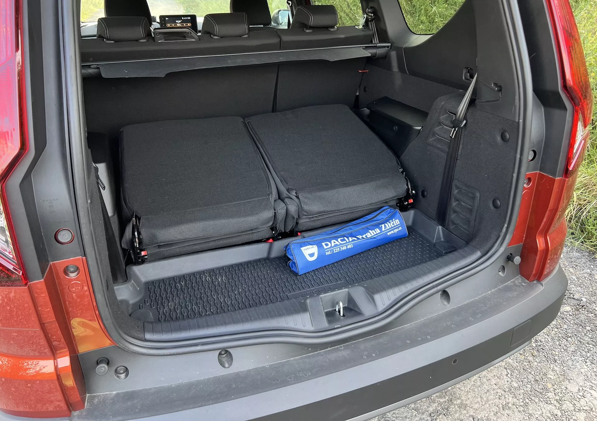 2022-es Dacia Jogger csomagtartójának részlete az nyitott rakodótérrel, összehajtott hátsó ülésekkel, tárgyak rögzítésére szolgáló textil hurkokkal, és egy kék Decaux hengerrel a szőnyegpadlón