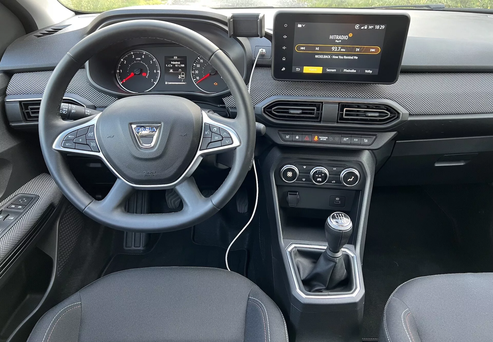 A 2022-es Dacia Jogger belső nézete a hátsó ülésről, kiemelve a sofőr műszerfalát, a gyártó logójával és vezérlőgombokkal ellátott kormánykereket, a digitális-analóg műszerfalat, az érintőképernyős infotainment rendszert, a HVAC vezérlőket, a kétzónás klímavezérlést és a kézi sebességváltót