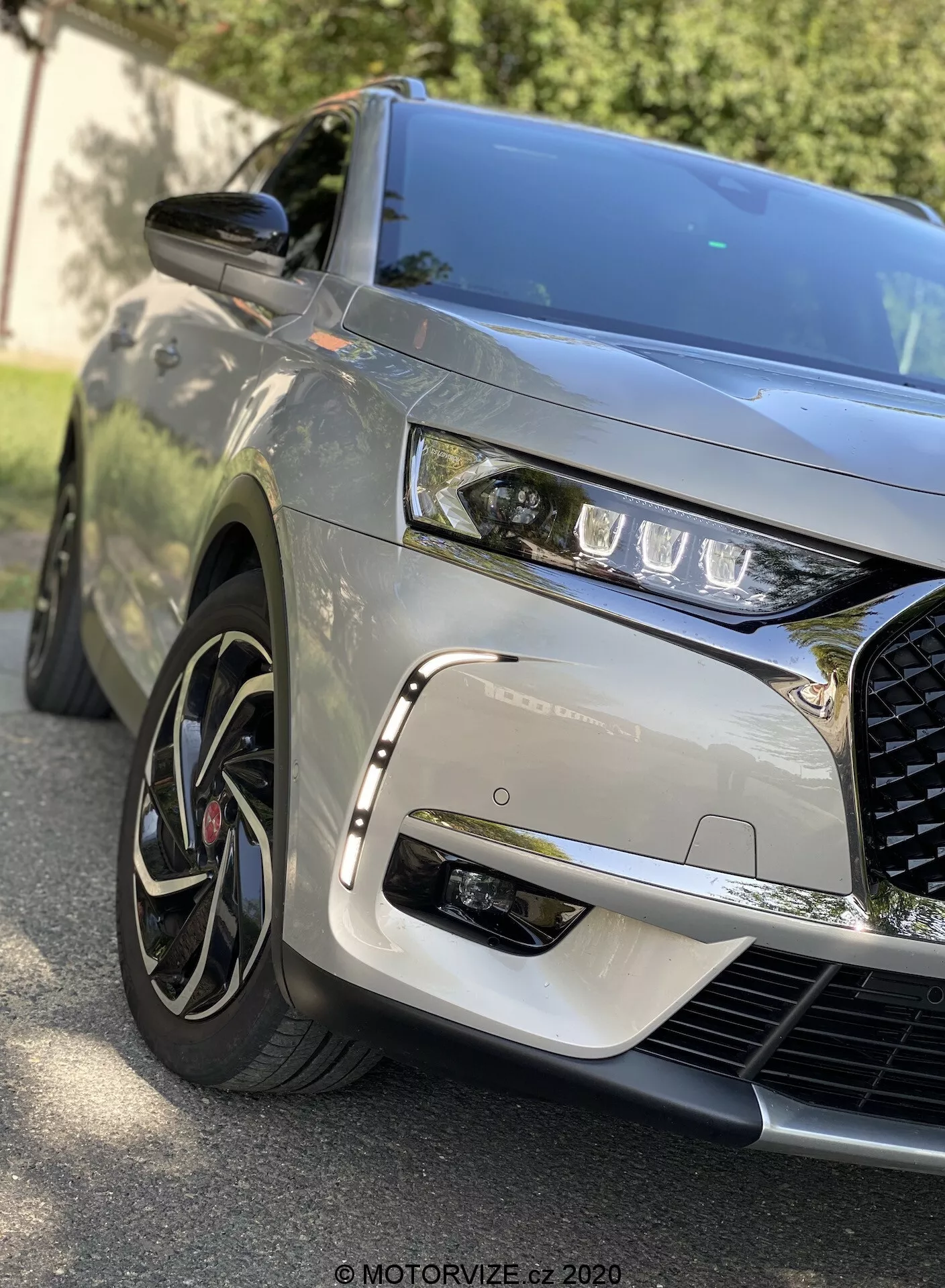 Közelkép előre néző szögből a 2018-as DS Automobiles DS 7 Crossback-ről, amely a bal első negyedet ábrázolja, elegáns LED fényszórókkal, kiemelkedő, bonyolult hűtőráccsal, szobrászvai motorháztetővel, többrácsos alumínium kerekekkel alacsony profilú gumikkal, metálfényű festéssel, test színű oldalsó tükörrel és dinamikus tükröződéssel, amely külső környezetben készült.