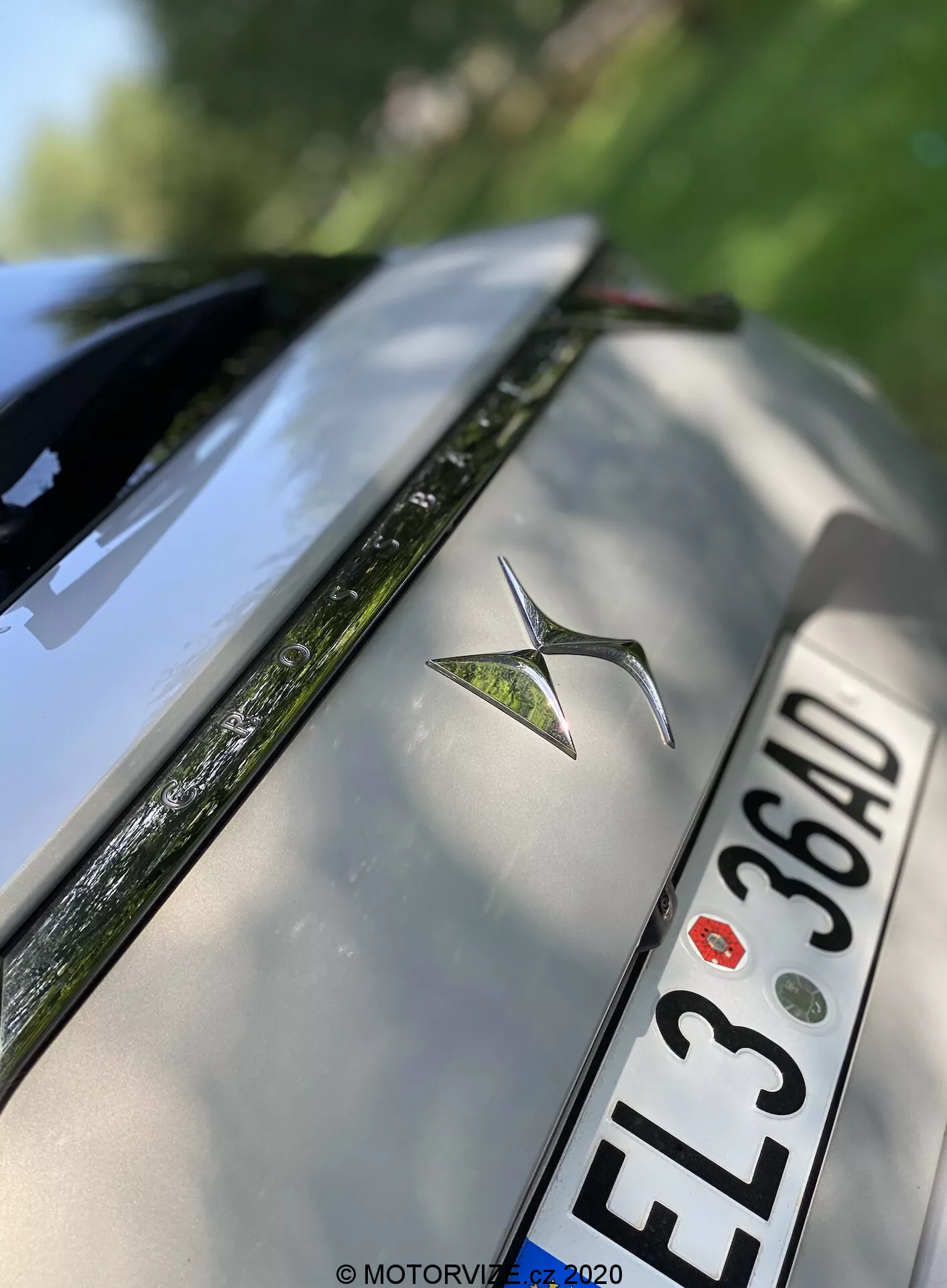 Az ezüst színű 2018-as DS Automobiles DS 7 Crossback hátsó részének szögletes közeli fotója. A kép kiemeli az autó fémből készült emblémáját a hátsó fedél közepén, az alatta lévő európai stílusú rendszámtáblát fekete karakterekkel 'EL3 36AD', és a lökhárító felső szélét. A hátsó szélvédő része a látható párátlanítóval és a magas fényű felületek tükröződései is láthatók. A sekély mélységélesség az emblémát hangsúlyozza, miközben a csomagtér fedelének és a hátsó szélvédő szélének elmosódásával lágyítja a képet.