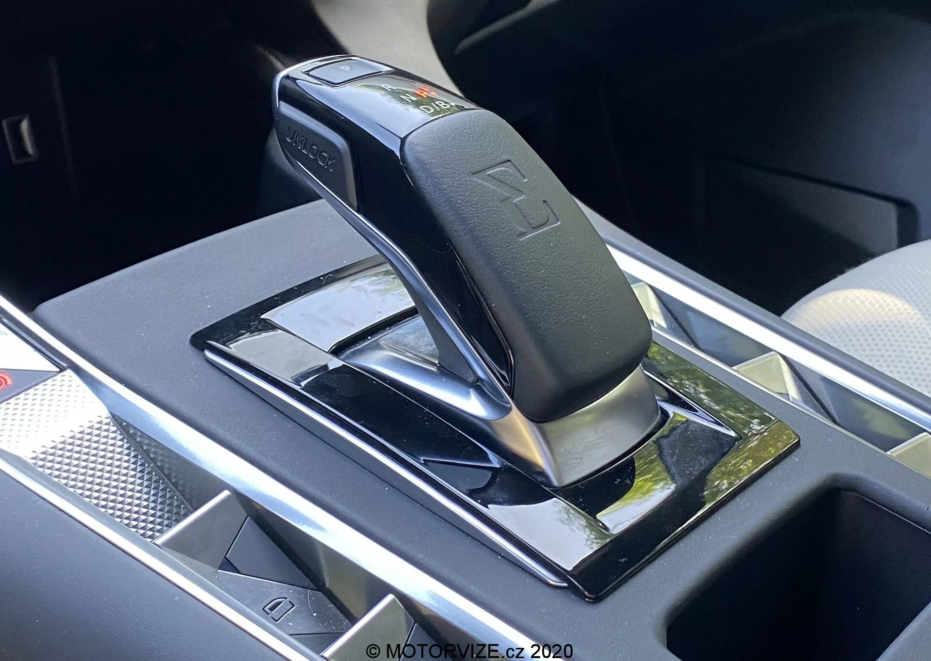 Közelkép az automata sebességváltó karjáról a DS Automobiles DS-7 2018 (DS-7 Crossback) modellnek, amely egy fényes fekete kart mutat ezüst díszítéssel, valószínűleg parkoló vagy üres helyzetben, fényes fekete panelre szerelve, fényvisszaverő felületekkel. A jellemzők közé tartoznak a sebességjelző szimbólumok, egy biztonsági gomb a karon, valamint egy tároló rekesz a közelben egy potenciális vezeték nélküli töltővel. Az ezt körülvevő terület krómozott vagy ezüst színű díszítéssel rendelkezik, ami modern, prémium járműbelsőt jelez.