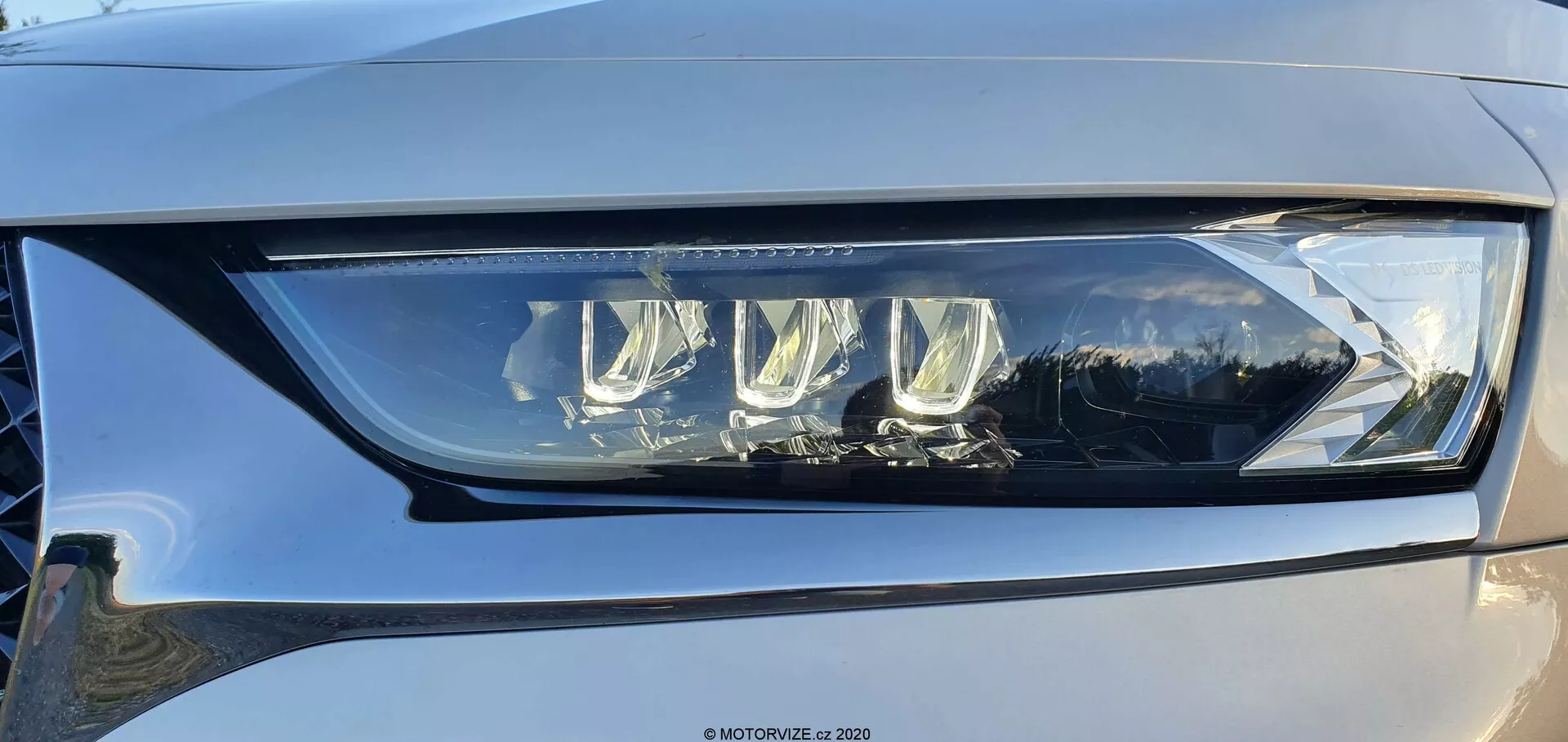 Közeli kép a DS Automobiles DS-7 Crossback bal fényszóróegységéről enyhe szögből, amely egy modern LED dizájnt, több vetítőlencsét és egy jellegzetes LED nappali menetfényt mutat, amely átlátszó, kiváló minőségű műanyagba van ágyazva. A fényszóró fejlett gépjármű világítástechnológiát példáz, amely képes alacsony és nagy fényerejű funkciókkal rendelkezni, potenciális adaptív jellemzőkkel a jobb láthatóság érdekében. Egy finom visszaverődést és egy kevés világos-közép szürke színt is láthatunk az autó felületén.