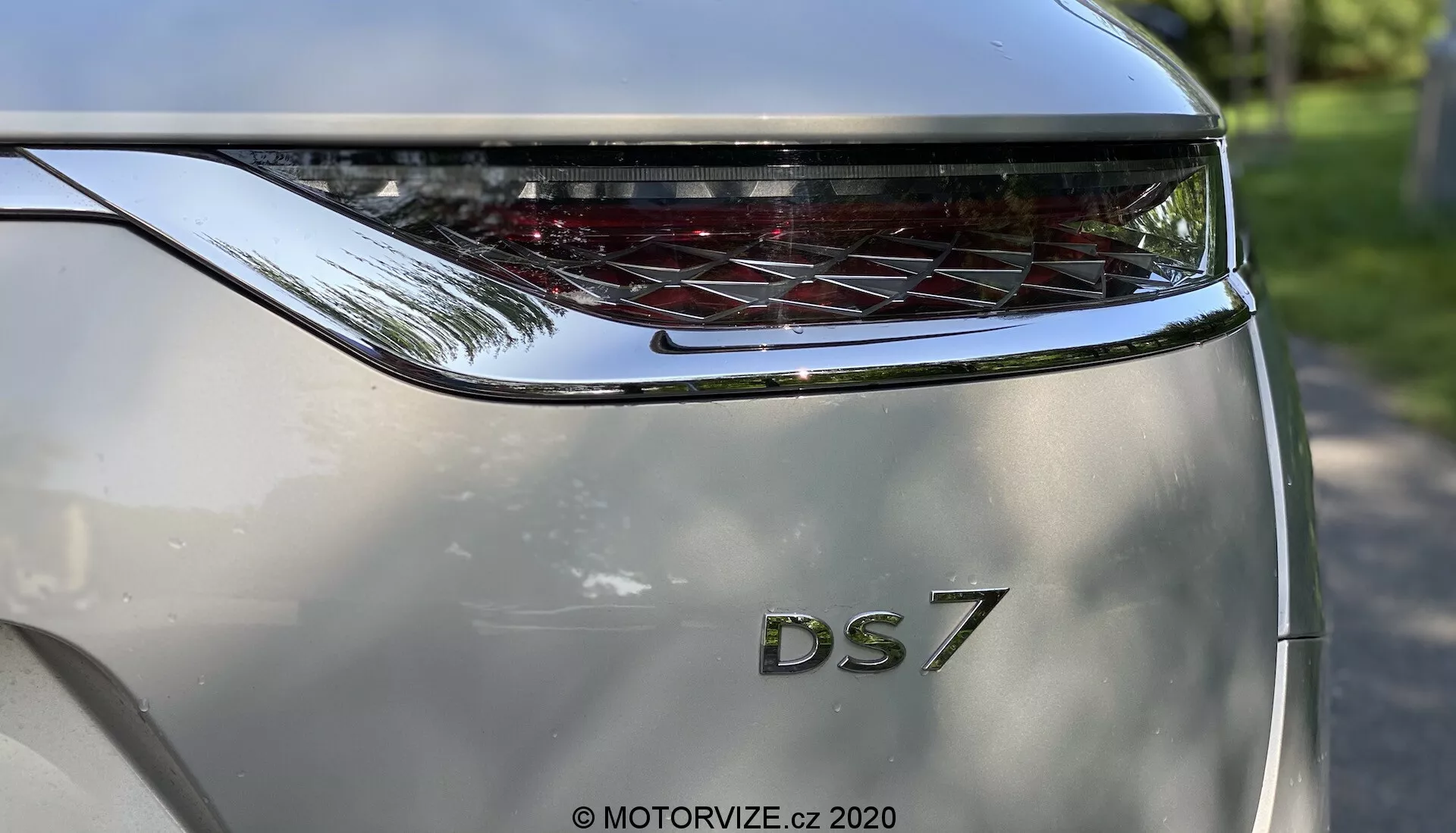 A DS Automobiles DS 7 Crossback 2018-as modell hátsó részének közeli nézete, alacsony szögből fényképezve. A fókusz a csomagtérajtón van, melyen feltűnő, vastag 'DS' felirat látható a '7' modellmegjelölés felett. A modellnév felett egy ívelt, vízszintesen húzódó hátsó lámpa található, jellegzetes LED elemekkel. Az autó fémes ezüst színe és a hásított dizájn modern és luxus megjelenést sugall. A fotó hangsúlyozza a márkajelzést és a hátsó lámpa dizájnját, miközben az autó felületén finoman tükröződik a fényes kültéri környezet.