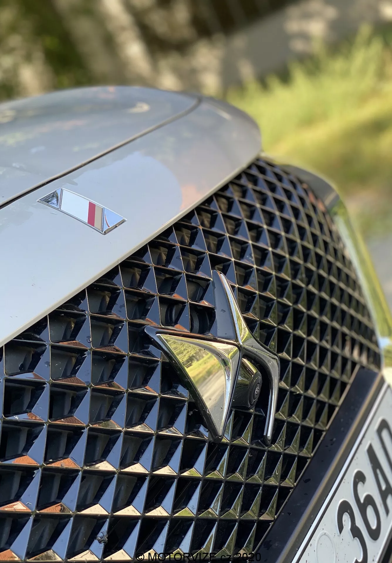 Közeli kép a 2018-as DS Automobiles DS 7 Crossback bal első részéről. A kép a fényűző, részletes, gyémánt mintázatú tükröződő felületekkel ellátott első hűtőrácsra összpontosít, középen egy hangsúlyos, stilizált logóval. A hűtőrács felett a motorháztetőn egy kisebb embléma látható, amely egy felső kategóriás modellt jelöl. A háttér homályos, kiemelve a hűtőrács megjelenését és a márka elemeit.