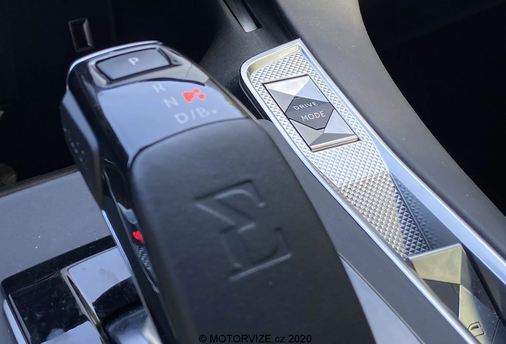 Közeli kép egy 2018-as DS 7 Crossback középkonzoljáról, amely a sebességváltót és a vezetési mód kapcsolót egy ferde szögből mutatja. Az L-alakú elektronikus sebességváltó kiemeli a parkoló pozíciót vörös LED fényjel és gombfunkcióval. Jobbra egy gyémánt mintás vezetési mód kapcsoló és egy ezüst berakás jelzik a prémium belsőt és a fejlett járművezérlési rendszereket.