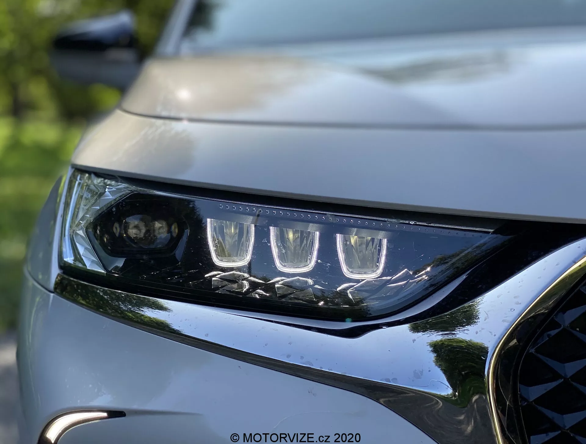 Közelkép a 2018-as DS Automobiles DS 7 Crossback bal első fényszórójáról és első hűtőrácsáról. A fényszóró jellegzetes LED nappali menetfényekkel és főszálhoz való projektorlencsével rendelkezik, több LED elemmel keretezve. A fényszóró elegáns és szögletes burkolata a modern aerodinamikát mutatja. A sötét, fényes hűtőrács egy része fémes részletekkel és a formás motorháztető egy része kiemeli a jármű dinamikus, prémium dizájnját. A háttér elmosódott, kiemelve az autó világítása és első részének éles és részletes formáit.