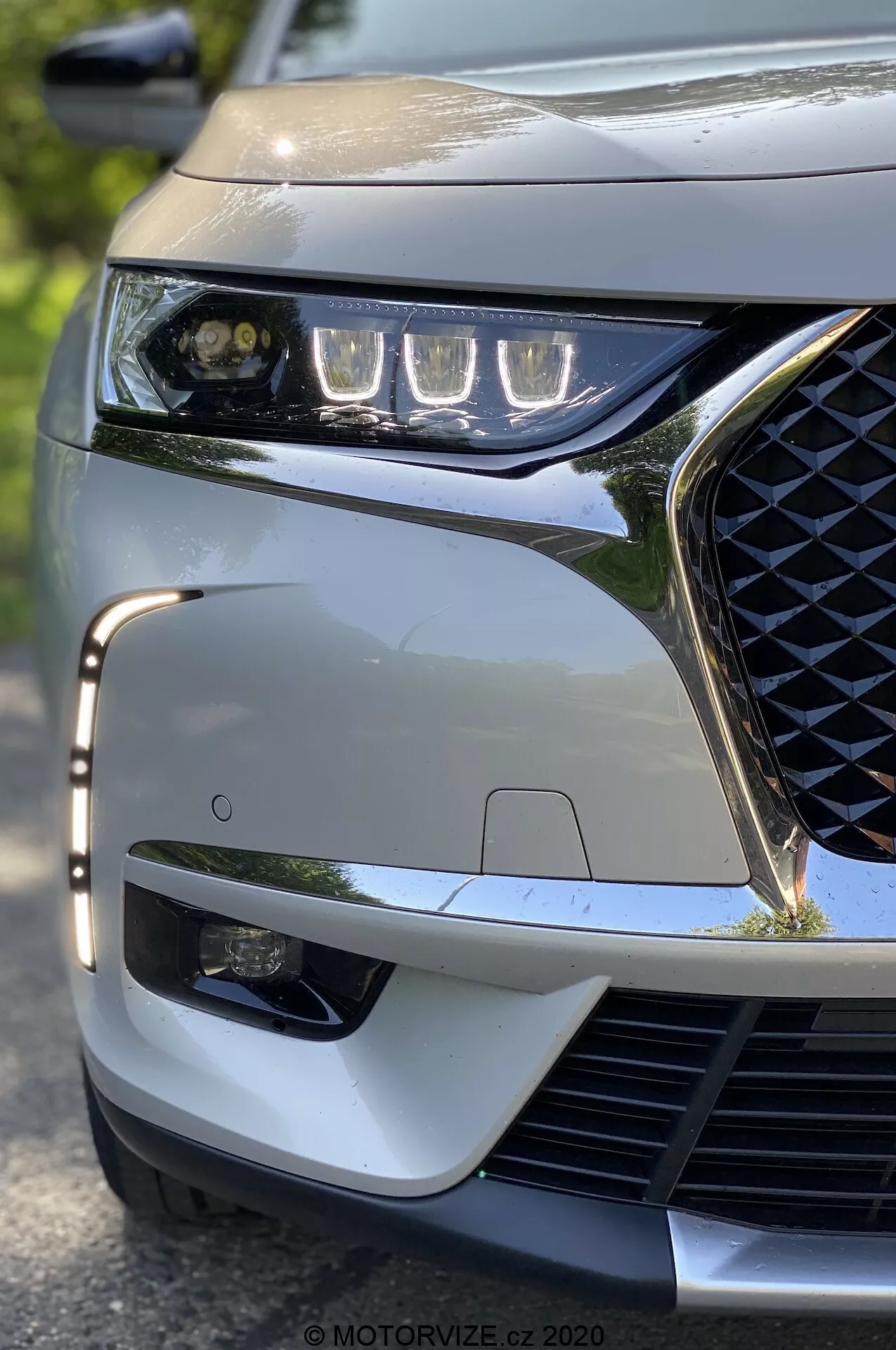 A 2018-as DS Automobiles DS 7 Crossback közelképe alacsony szögből, a bal első sarokra fókuszálva. Kiemelésre kerül a LED-es fényszórók elegáns kialakítása, a geometrikus mintázatú hűtőrács, a sportos elülső lökhárító és a látható ADAS érzékelő, amelyek a jármű modern technológiai és esztétikai jegyeit hangsúlyozzák.