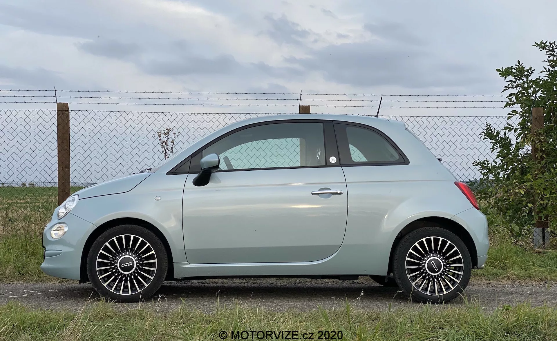 Modern szubkompakt ferdehátú autó oldalnézeti szögből, amely a jármű teljes hosszát mutatja. Egy sík felületre parkolva látható, háttérben kerítéssel és felhős égbolttal. Jellemzői: oldalsó ajtók, ablakok, tükrök, kétszeres ajtó elrendezés, kis hátsó negyedablak, lekerekített tető, pasztell színű karosszéria kontrasztos tetővel, többrudas alufelnik, jó állapotú gumik, részletes dizájnvonalak és természetes nappali fény.