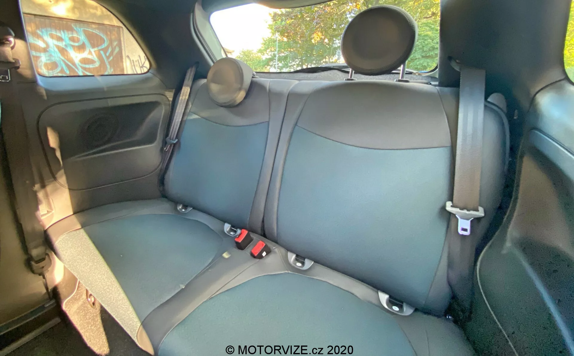 A 2015-ös Fiat 500 belseje látható a képen, amely a hátsó utasüléseket mutatja be bőr vagy műbőr kárpitozással sötét színben, látható varrással, hárompontos biztonsági övekkel, állítható fejtámlákkal, ajtópanellel elektromos ablakvezérléssel, tároló rekesszel, hangszóróráccsal, részleges kilátással a plafonra és felső kapaszkodóval. A kamera szöge kissé megemelt, elölről hátrafelé nézve, részletes nézetet biztosítva a hátsó padülésre, a hátsó ajtóra, a plafon egy részére és az ablakokra.